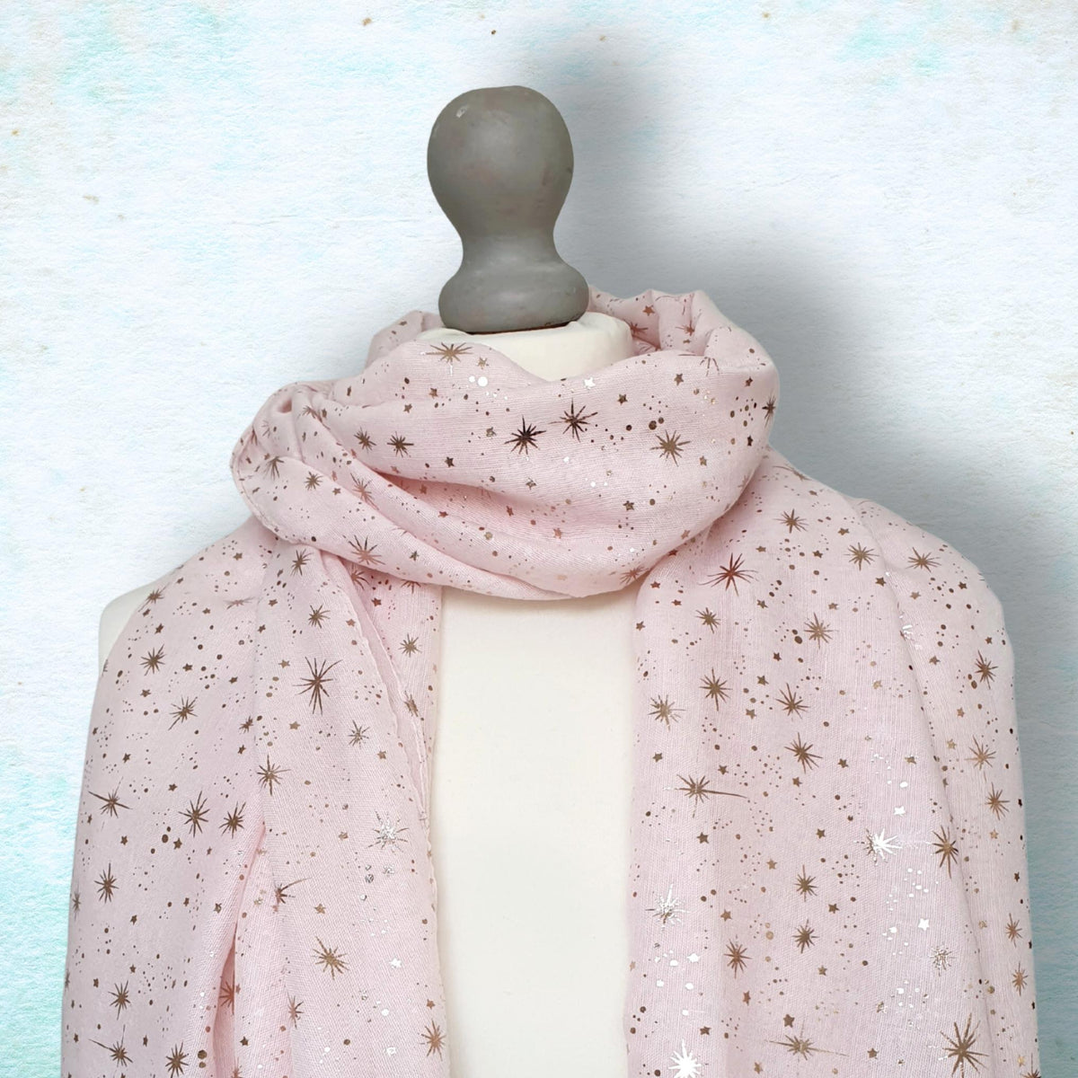 A light pink scarf with a metallic gold star pattern draped on a white mannequin torso against a textured light blue background.