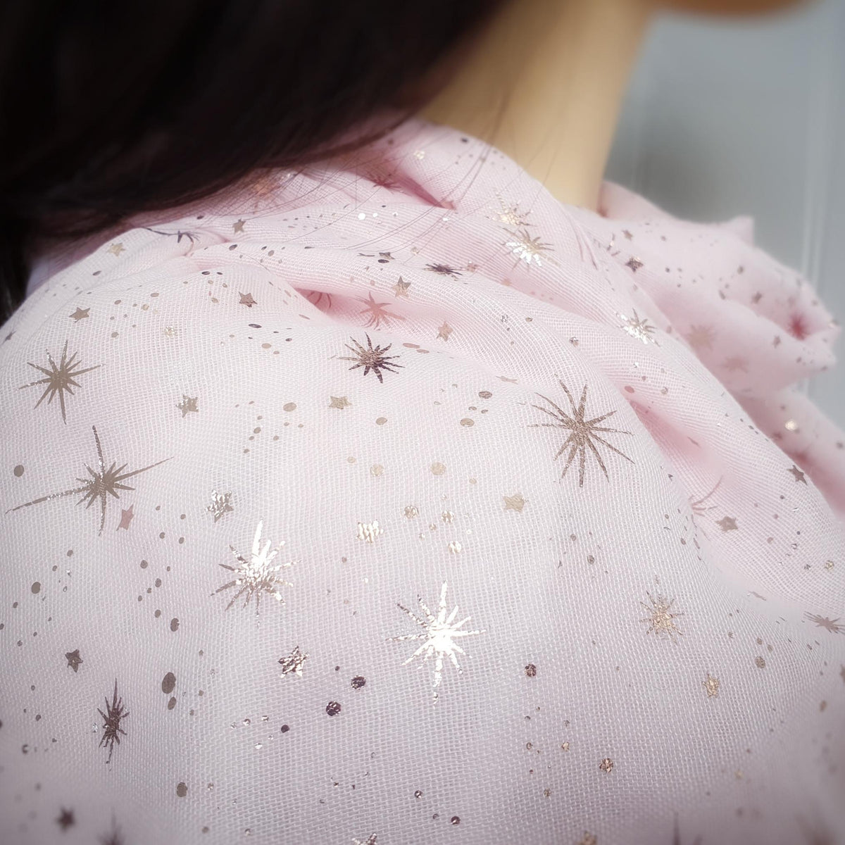 A close-up of a person wearing a light pink fabric scarf or shawl, decorated with metallic rose gold star and dot patterns.