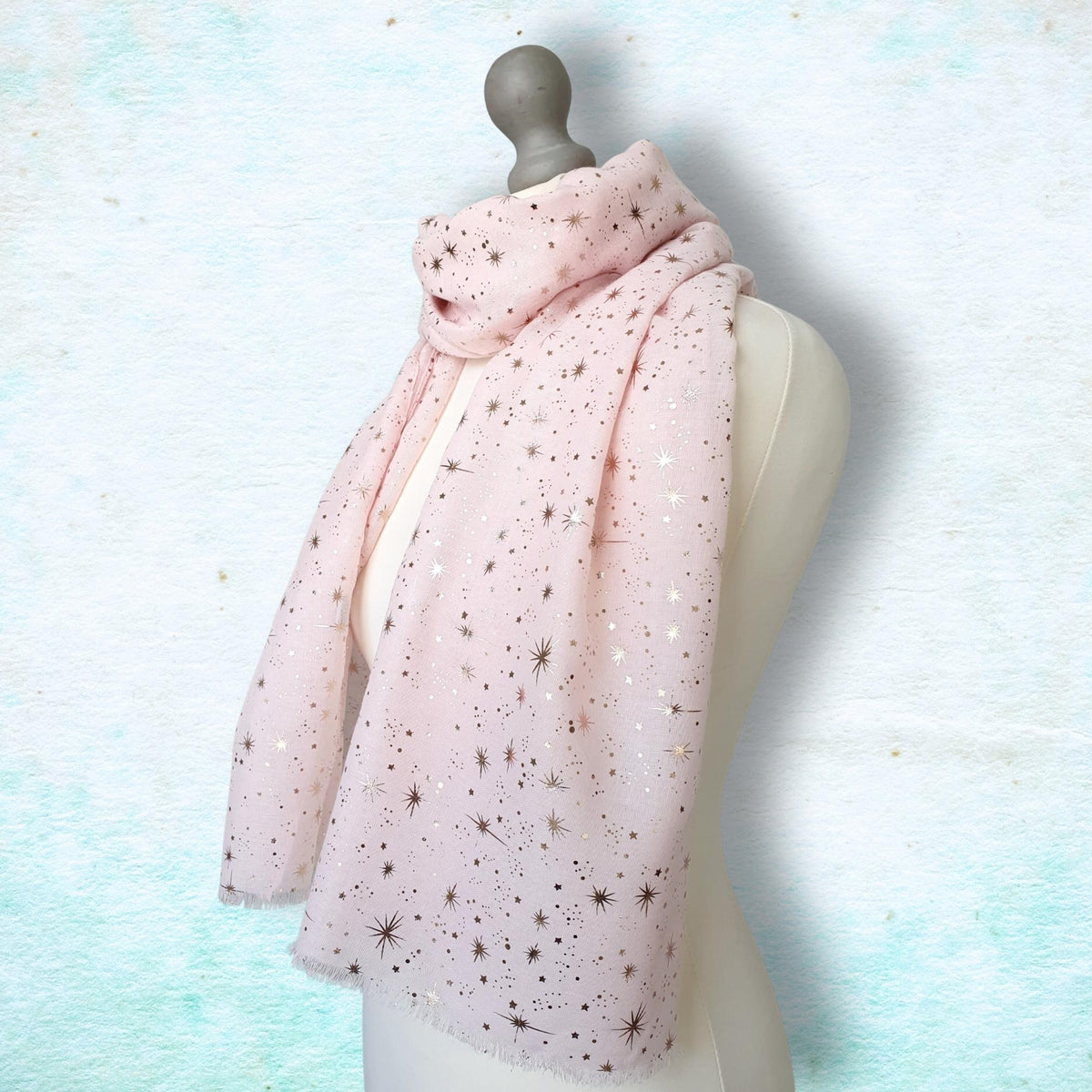A light pink scarf with small metallic star embellishments, draped on a cream-colored mannequin torso against a textured light blue and white background.