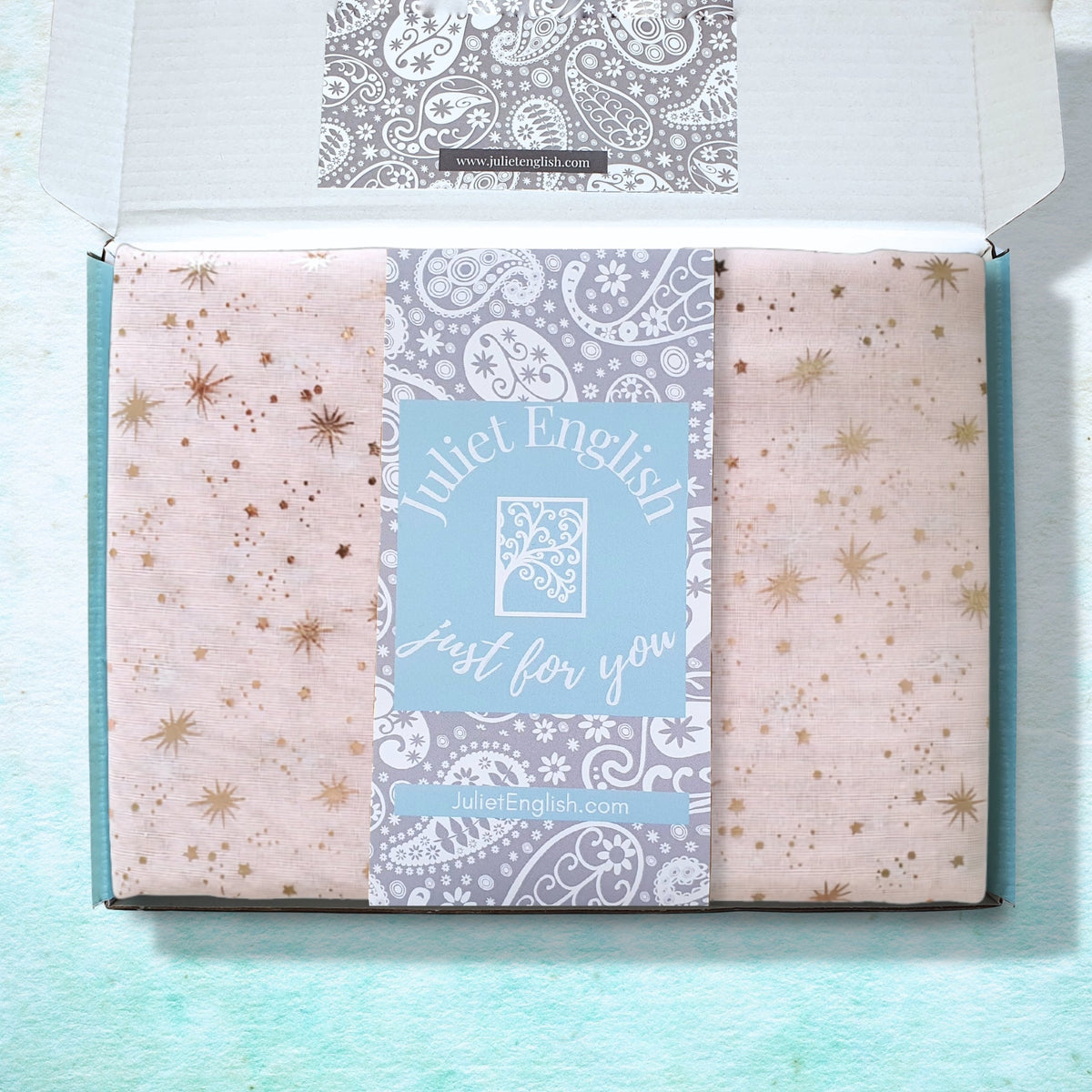 An open gift box containing a pale pink celestial patterned scarf with metallic gold star prints and a central light blue paper label reading 'Juliet English just for you' on a textured light aqua background.