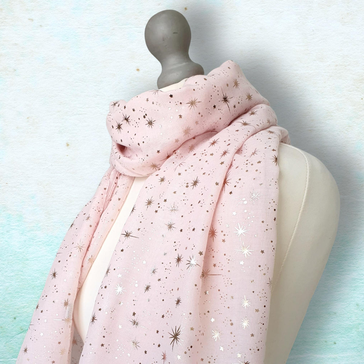 A light pink scarf with a scattered metallic gold star and sparkle pattern, draped on a cream-colored dress form mannequin against a textured light blue background.