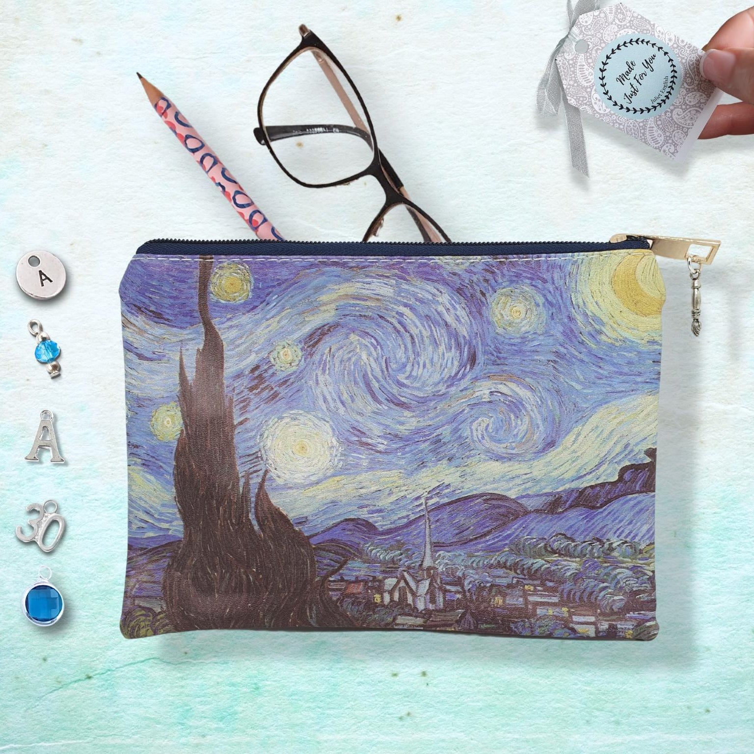 A zippered pouch featuring a printed Van Gogh-inspired night sky artwork in swirling blue, purple, and yellow tones. The pouch is shown on a pale textured background with styling props including eyeglasses, a pencil, small jewelry pieces, and a hand holding a small tag.