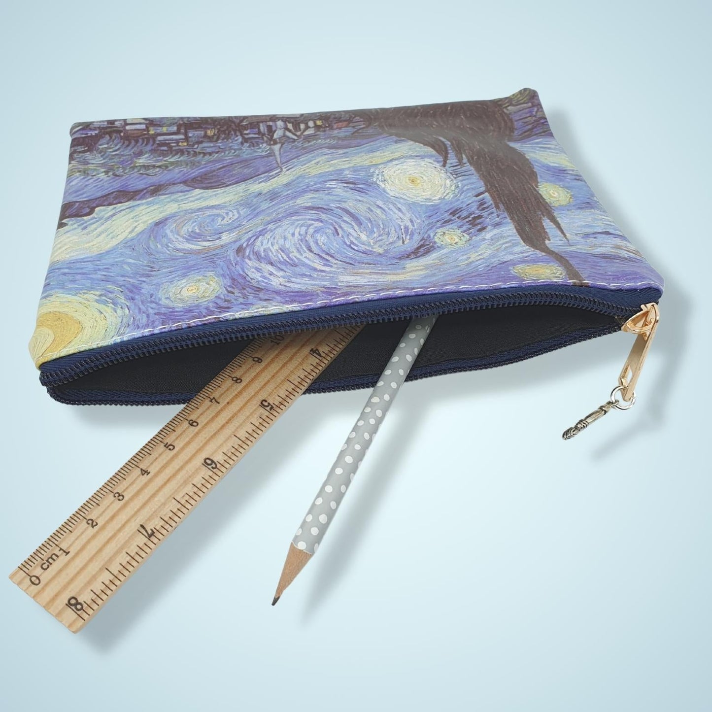 A zippered pouch featuring a print of Van Gogh's Starry Night, shown with a wooden ruler and a white polka-dot pencil protruding from the front, on a light blue background.