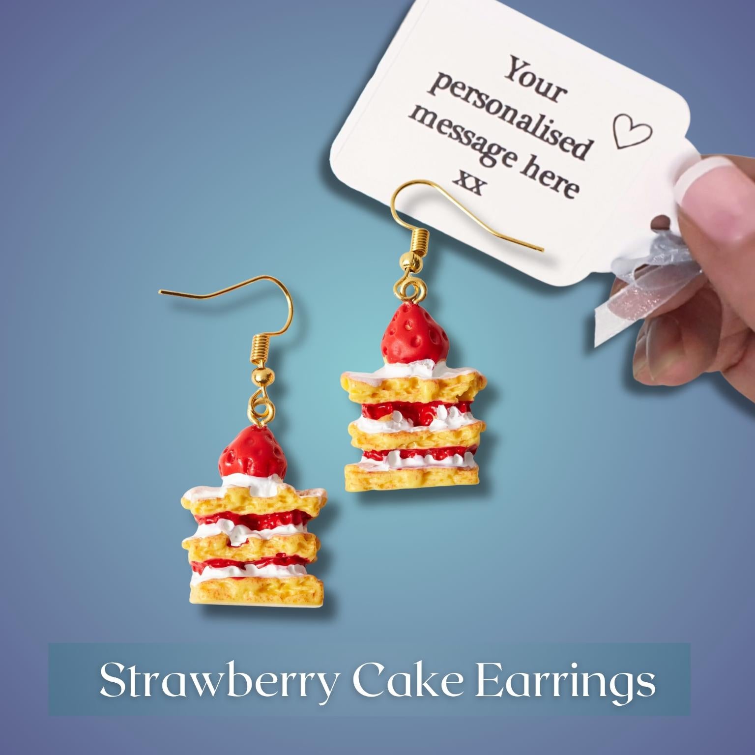 Strawberry cake earrings with gold-tone hooks, featuring red strawberry layers, white cream, and yellow cake layers, shown alongside a white sample tag and a hand holding the tag on a blue gradient background.
