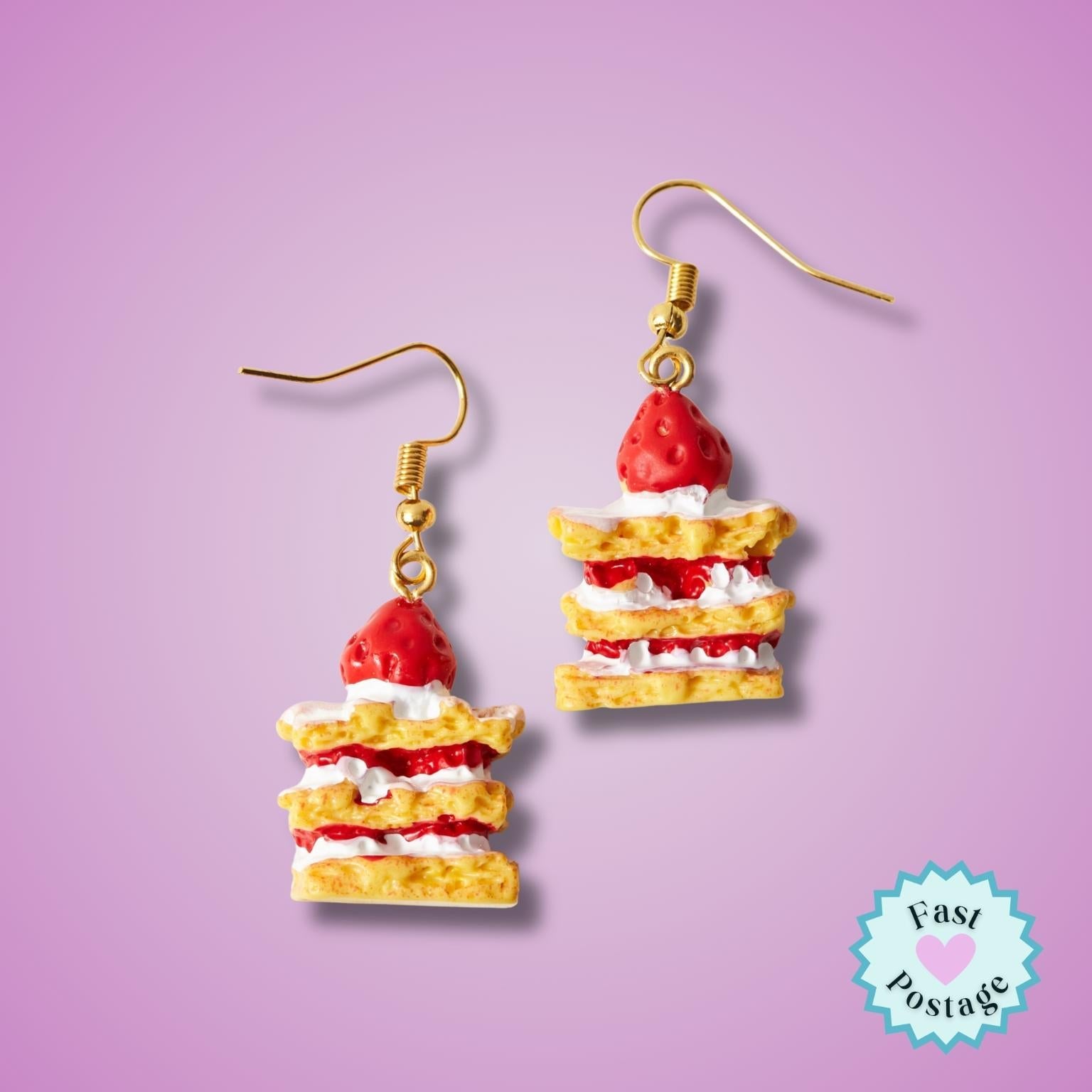 A pair of novelty dangle earrings shaped like strawberry shortcake slices, featuring red strawberries, white cream, and golden-brown cake layers, suspended from gold fishhook wires on a light purple background. A light blue 'Fast Postage' logo sticker is in the bottom right corner.