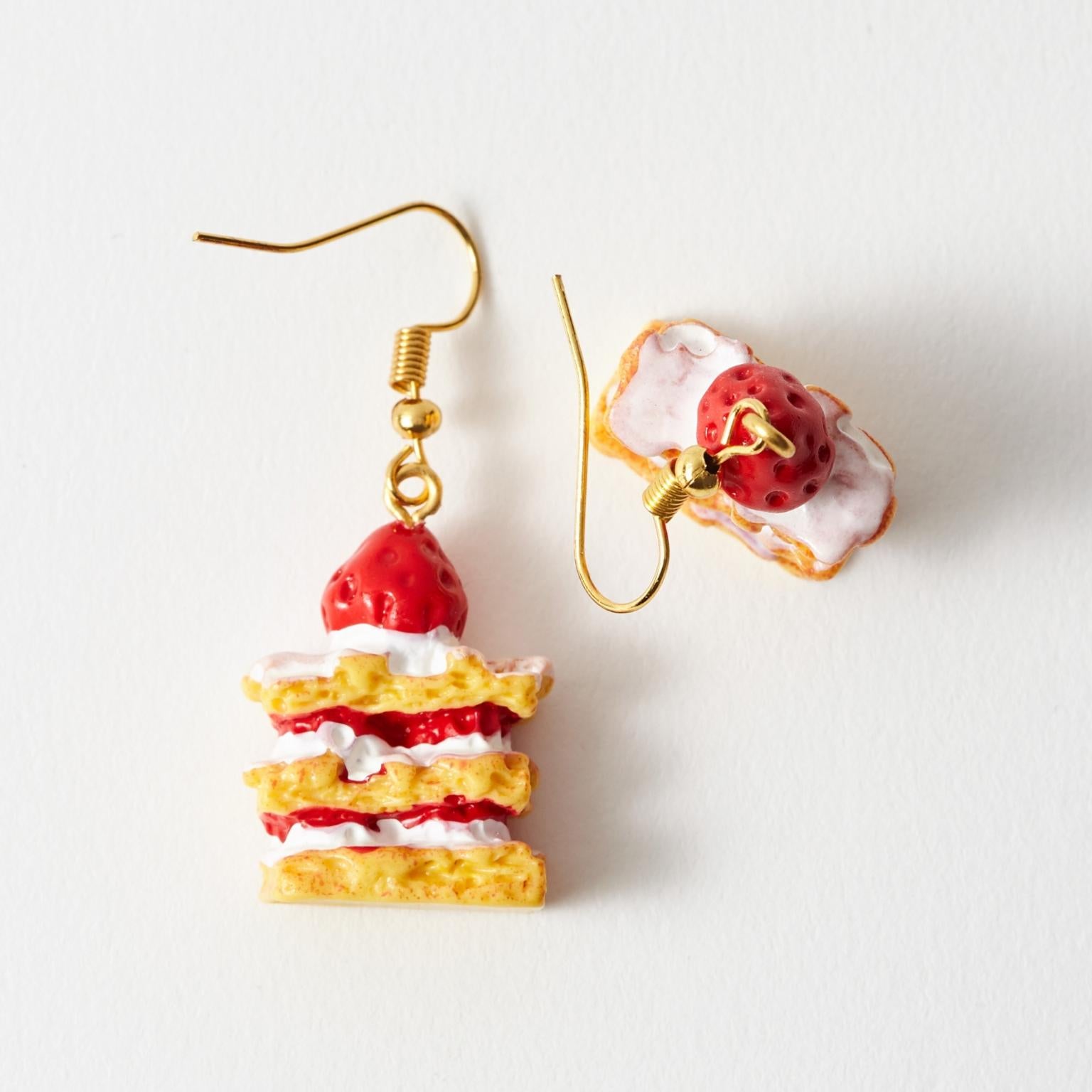 A pair of novelty drop earrings on a plain white background. One earring is shaped like a strawberry shortcake with gold-tone hooks, and the other is a matching close-up detail of the same design.