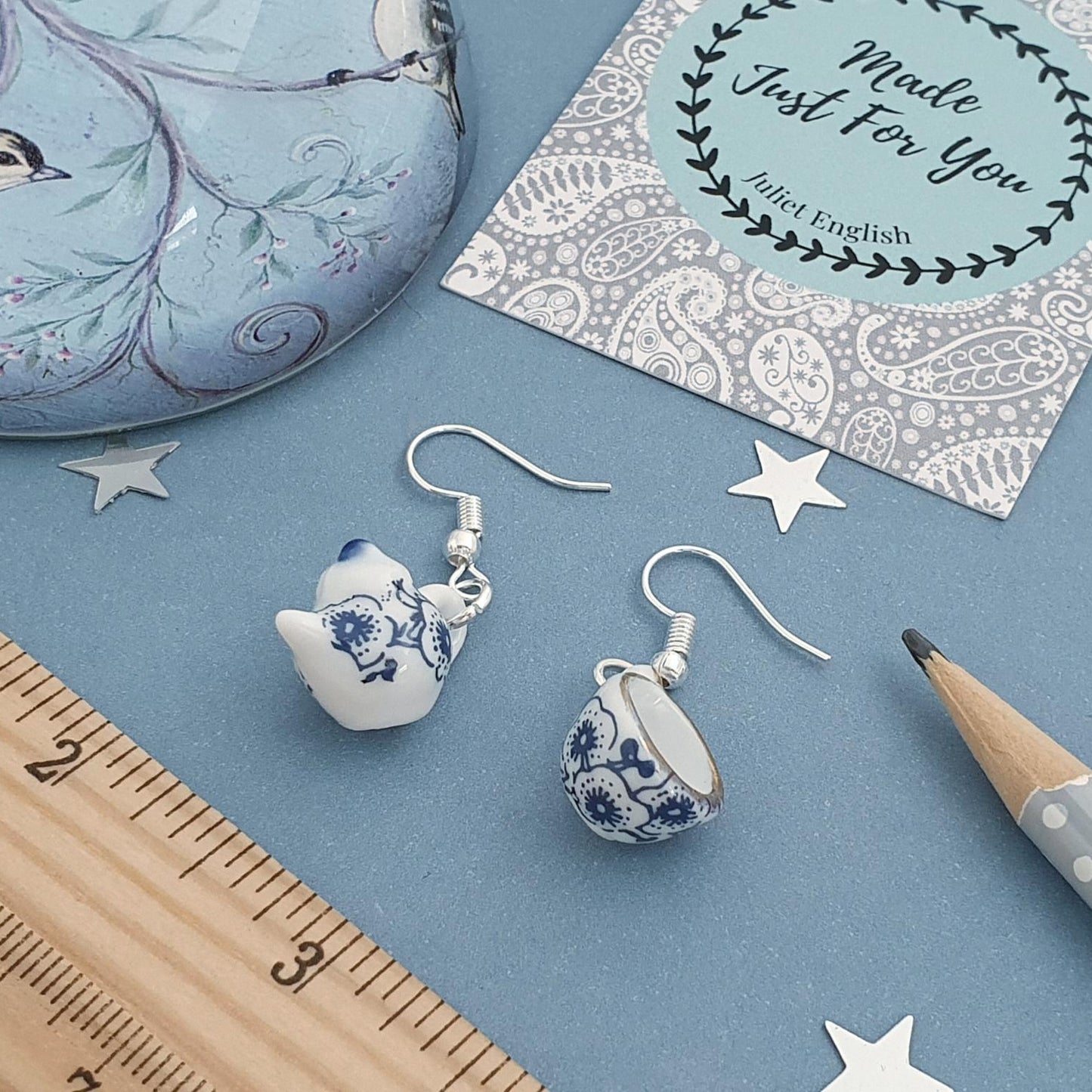 Tea cup and tea pot earrings on a blue surface with a ruler and card