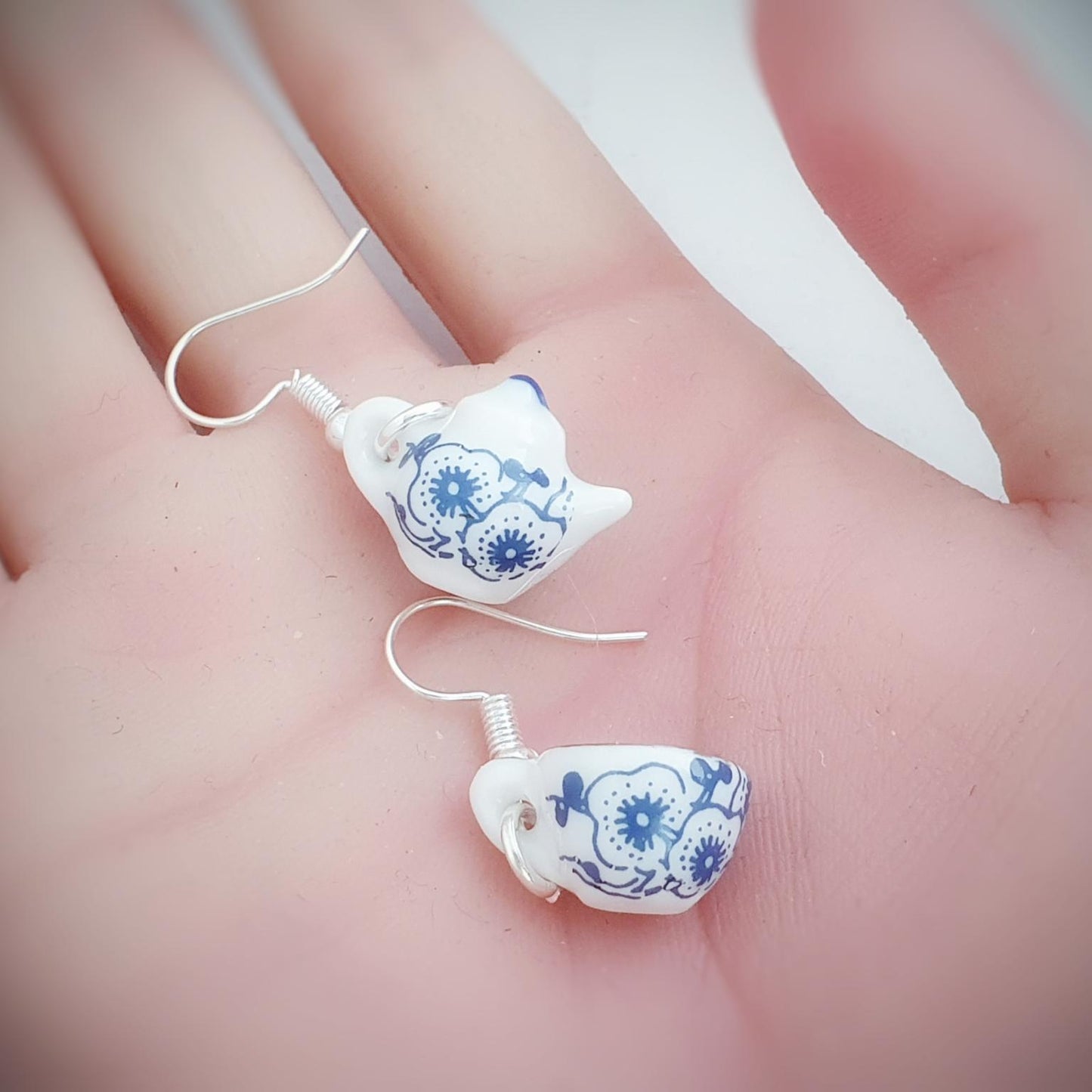 White ceramic teapot and tea cup earrings with blue floral patterns held in a hand against a blurred background