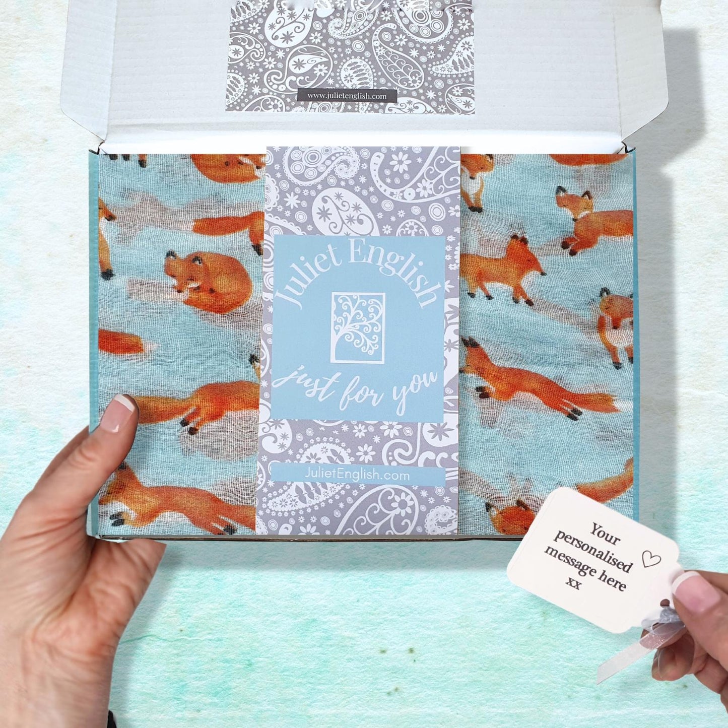 A person's hands holding an open gift box containing a folded patterned fabric scarf with light aqua blue background with orange fox illustrations, with a central label reading 'Juliet English just for you'. A small white card is shown in the bottom right corner.