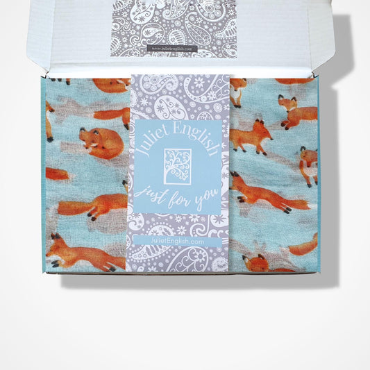 An open gift box containing a folded patterned fabric scarf with light aqua blue background with orange fox illustrations, with a central label reading 'Juliet English just for you'.