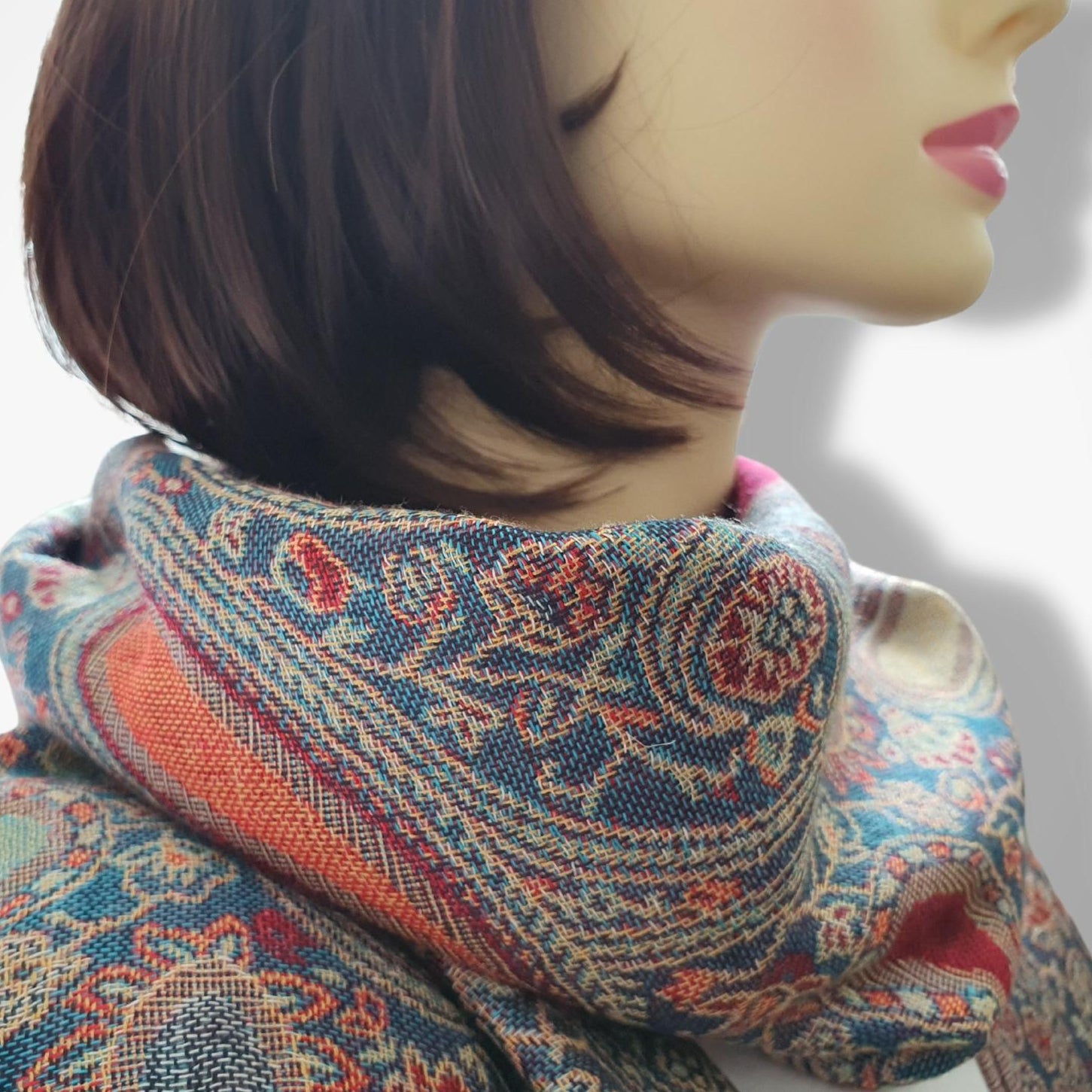 A close-up of a mannequin head with short dark brown hair wearing a patterned scarf in blue, rust orange, beige, and green tones against a plain light background.