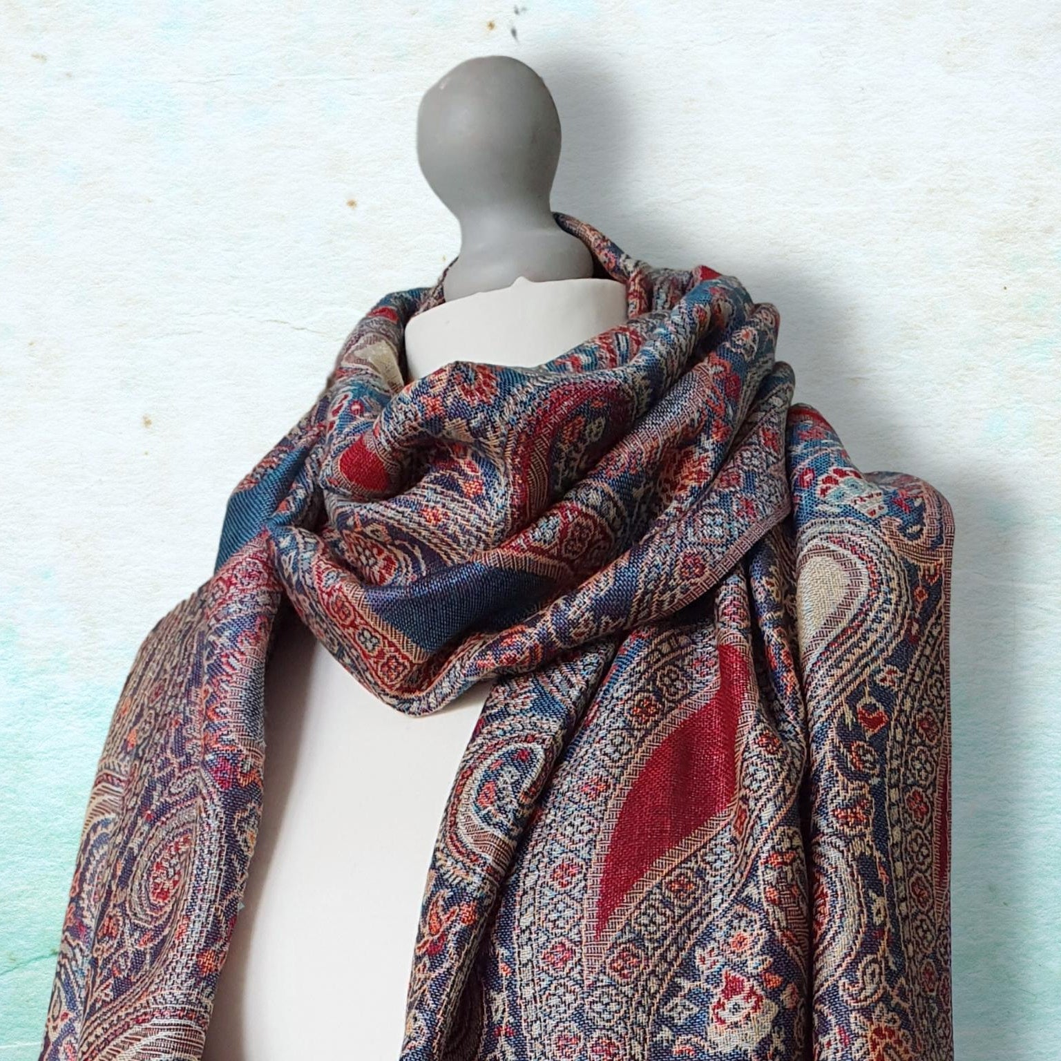 A multicolor paisley patterned scarf draped on a white mannequin torso against a textured light blue background.