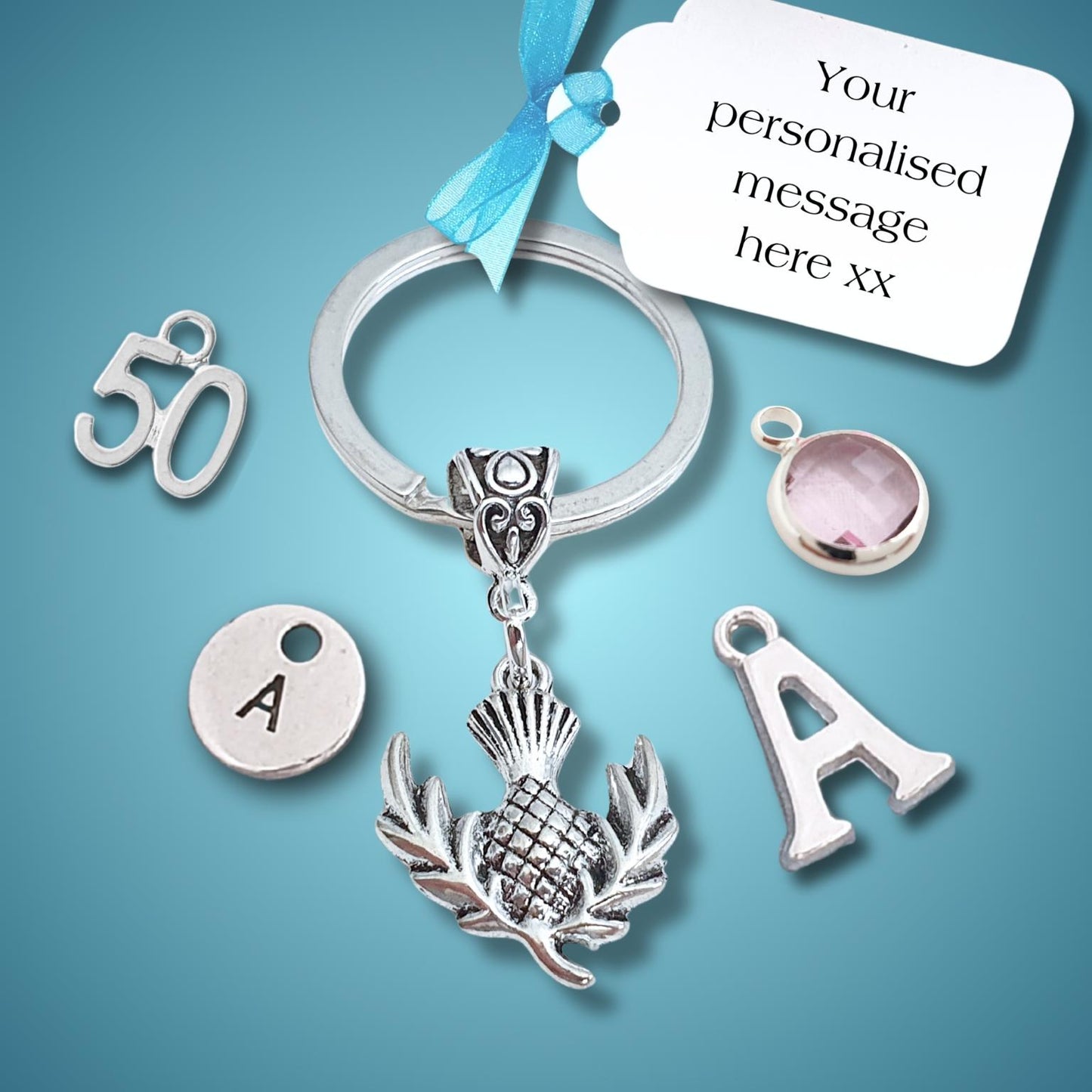 A silver keychain with a thistle charm, shown with several customizable charms on a blue gradient background: a '50' charm, a round silver disc stamped with 'A', a letter 'A' charm, and a pink gemstone bezel. A white tag with a blue ribbon reads 'Your personalised message here xx'.