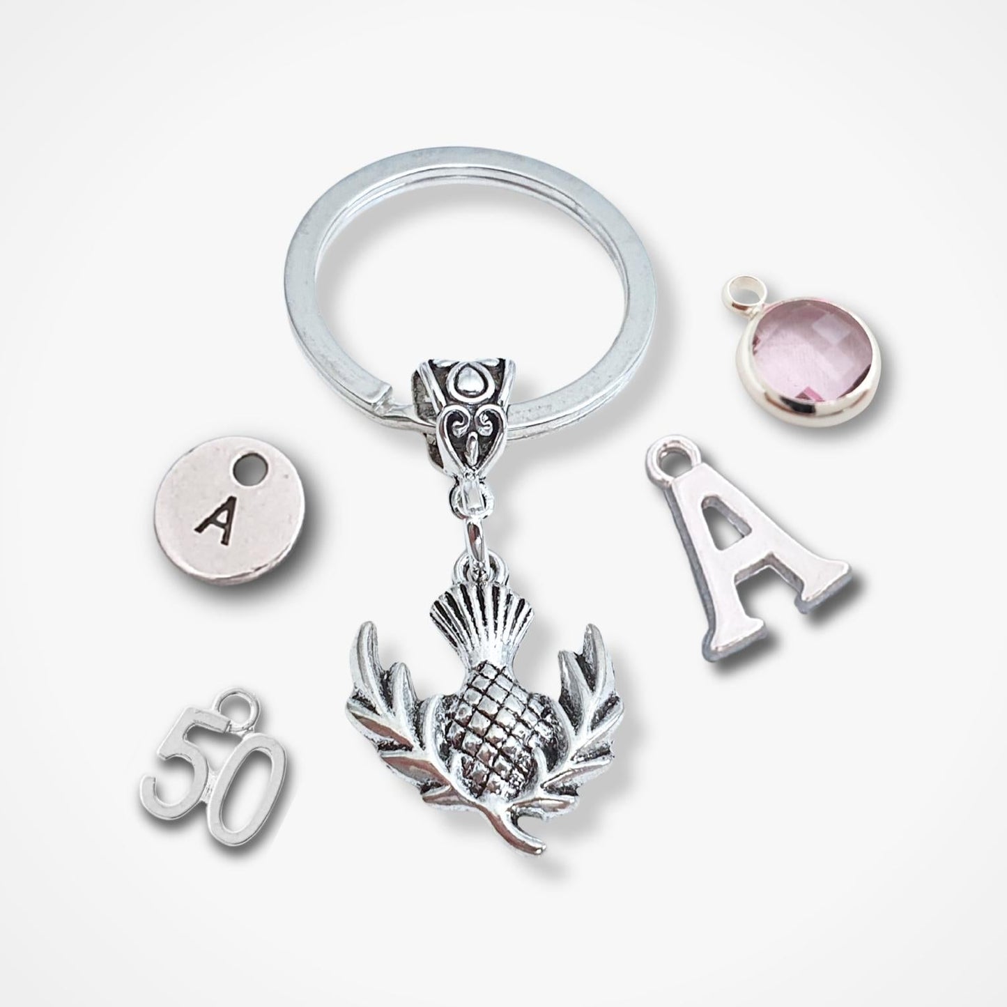 A silver keychain with a split ring and multiple charms, including a central thistle design, an 'A' letter charm, a '50' charm, and a round pink gemstone charm, displayed on a light gray background.