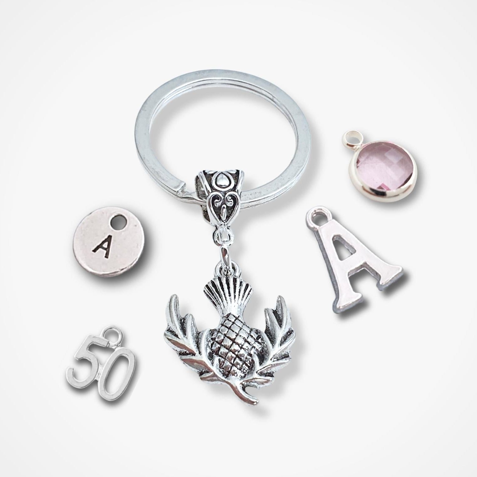 A silver keychain with a split ring and multiple charms, including a central thistle design, an 'A' letter charm, a '50' charm, and a round pink gemstone charm, displayed on a light gray background.