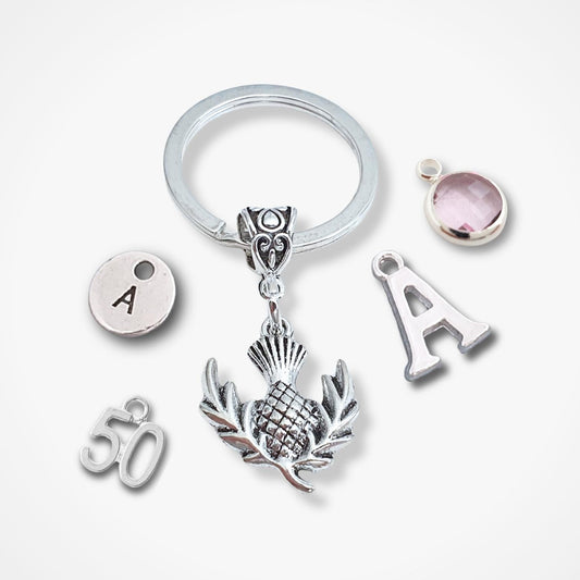A silver keychain with a split ring and multiple charms, including a central thistle design, an 'A' letter charm, a '50' charm, and a round pink gemstone charm, displayed on a light gray background.