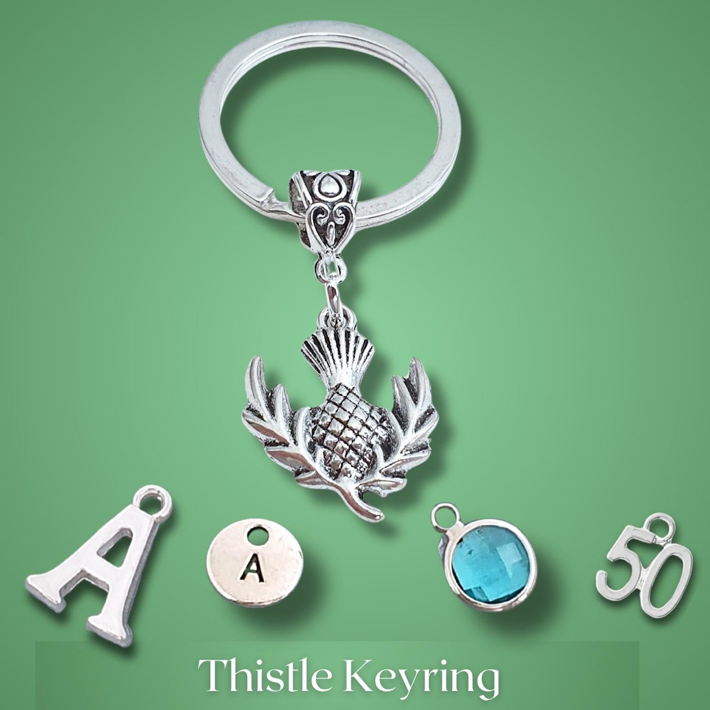 A silver keyring with a detailed thistle  charm, displayed alongside five separate silver charms (letter A, round charm with 'A' inside, a round blue gemstone bead, and the number 50) on a solid green background.