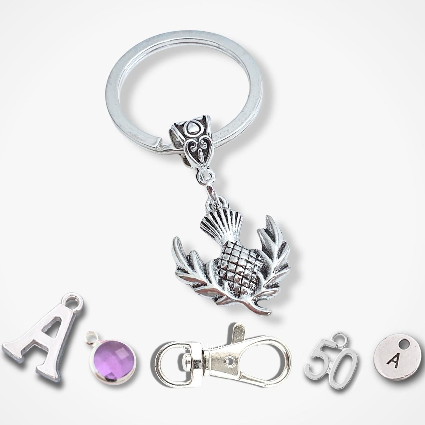 A silver metal keychain set featuring a detailed bail bead and thistle charm, a split key ring, a lobster clasp, and several small silver beads and charms, including a purple gemstone bead and a round bead with the letter 'A'.