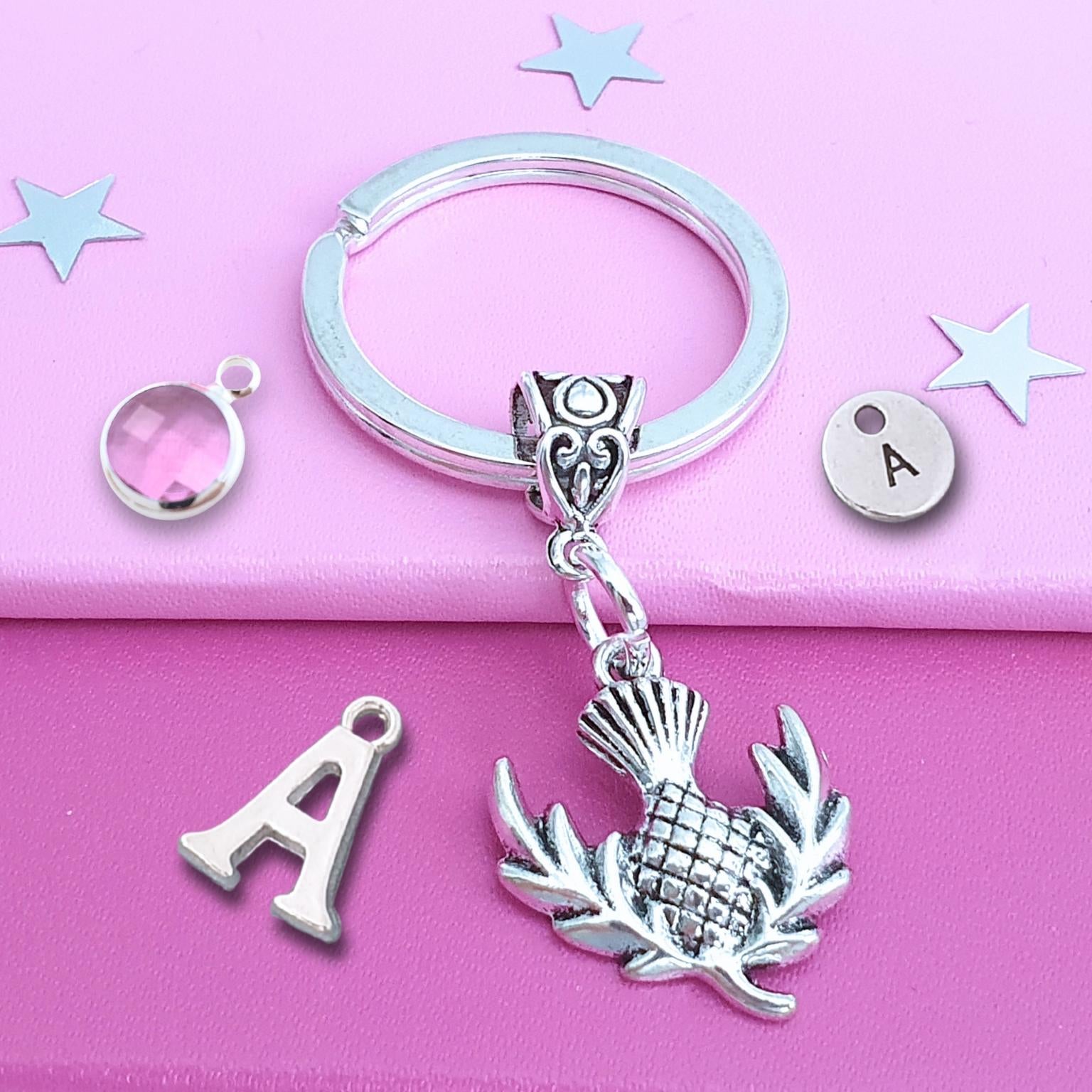 A silver keychain with a fancy bail bead and a thistle pendant, displayed on a pink background with matching silver letter 'A' charms and small decorative stars.