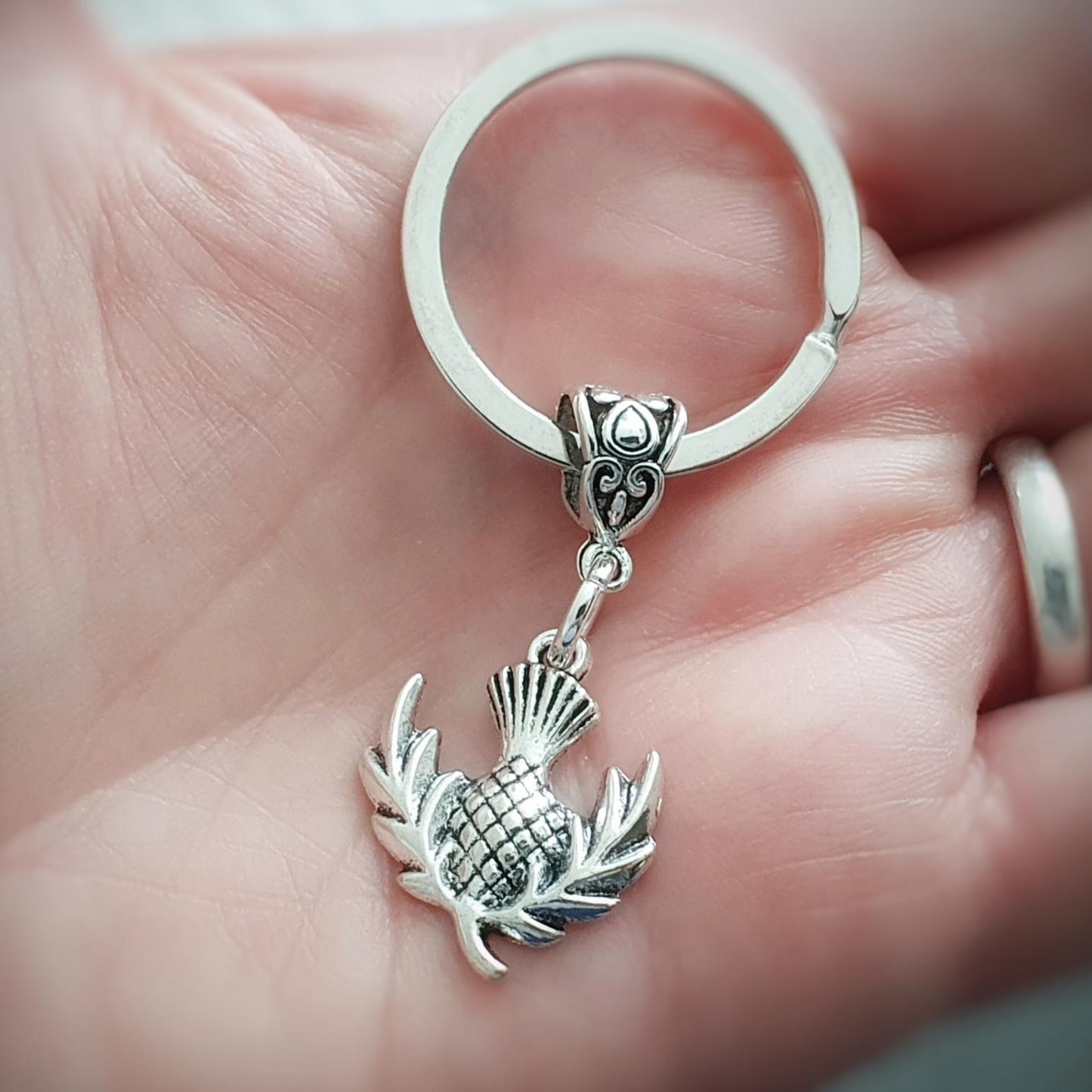 A silver metal key ring with a fancy bail bead and a dangling Scottish thistle charm, held in a person's hand.