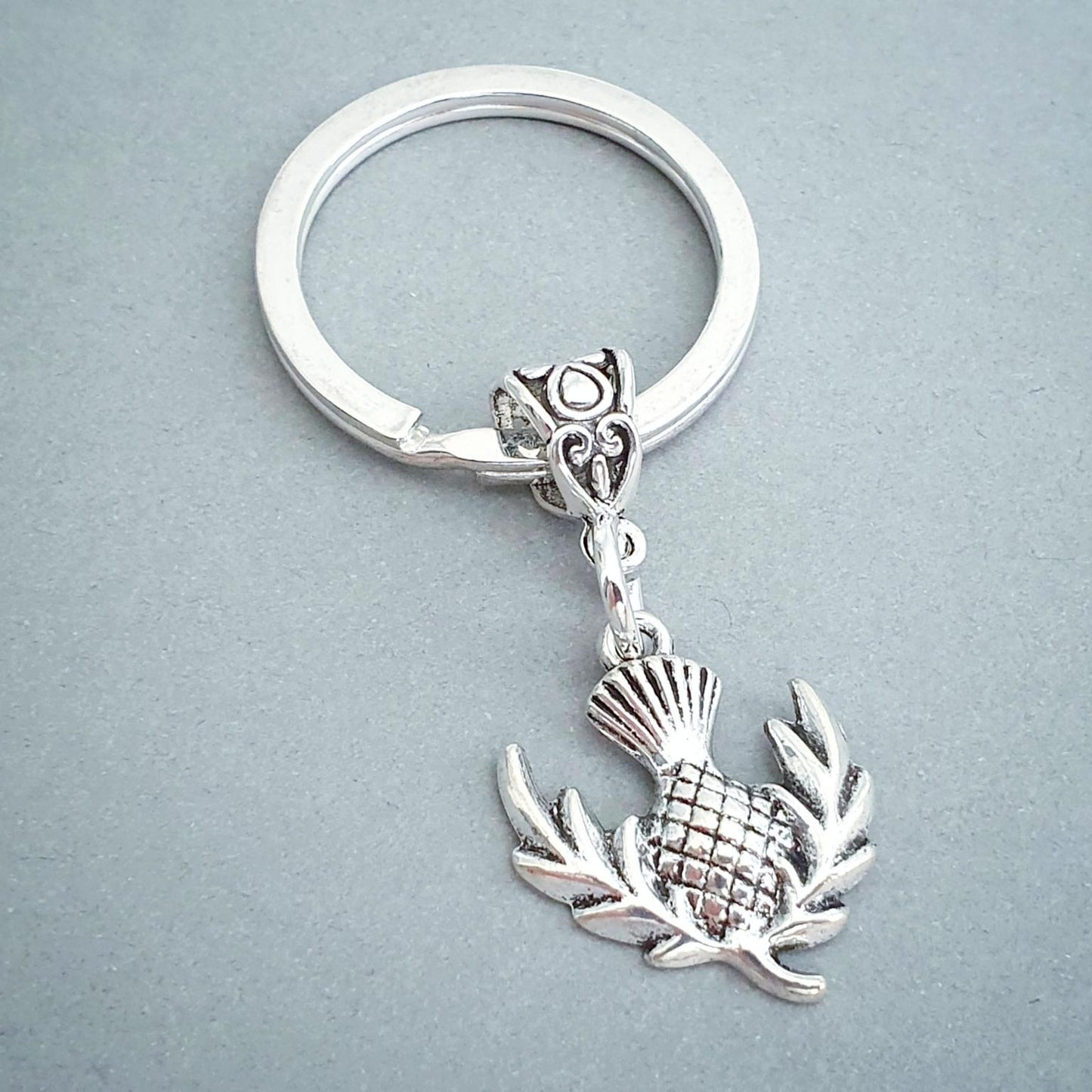 A silver metal keychain featuring a small thistle charm with leafy branches, attached to a split key ring, shown on a plain gray background.