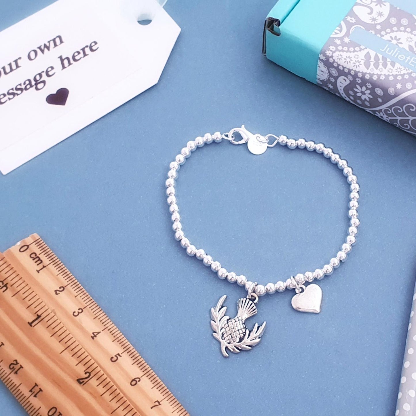 A silver beaded bracelet with a thistle and heart charm, displayed on a blue background with a wooden ruler, pencil, and gift box.