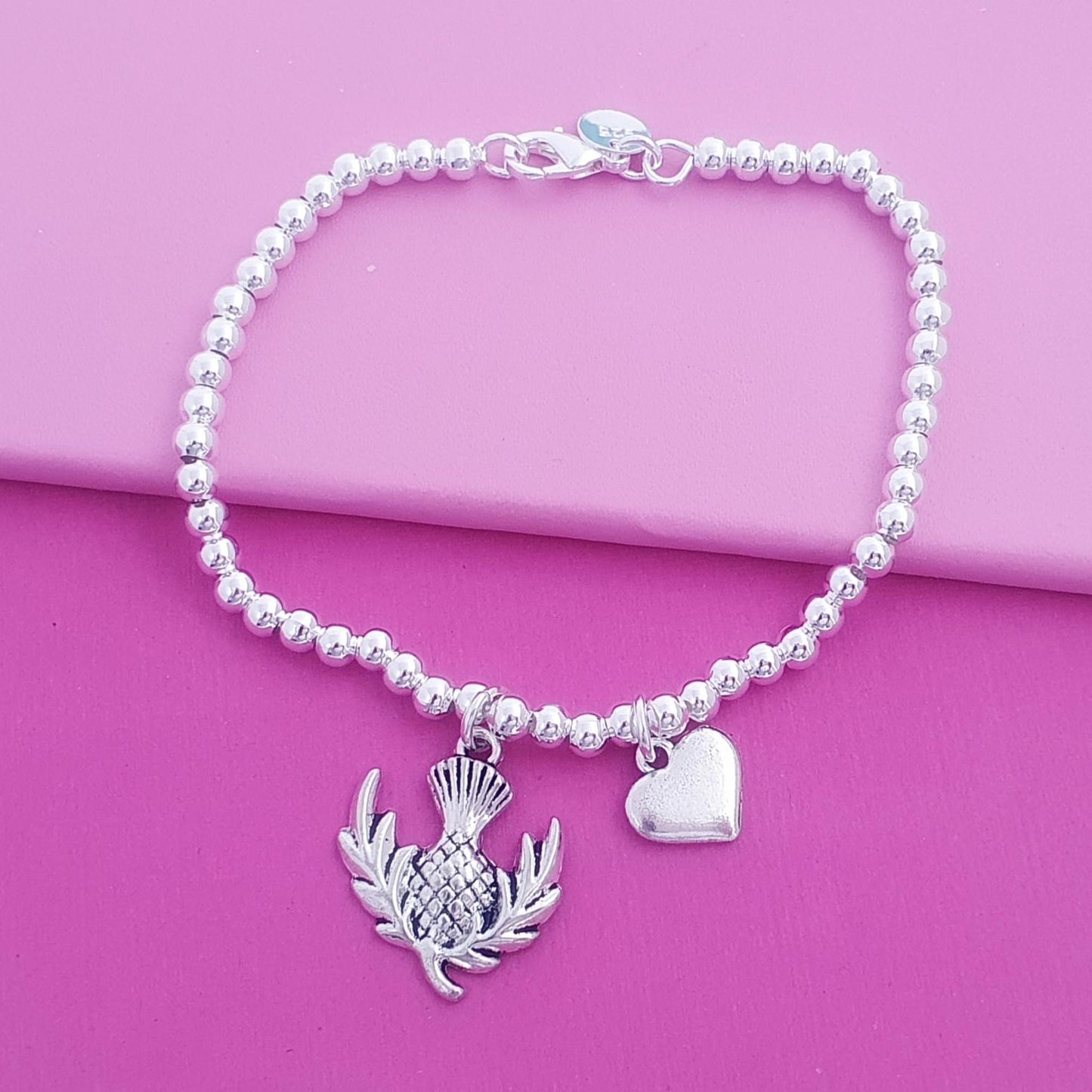 A silver beaded bracelet featuring a thistle and heart charm, displayed on a vibrant pink background.