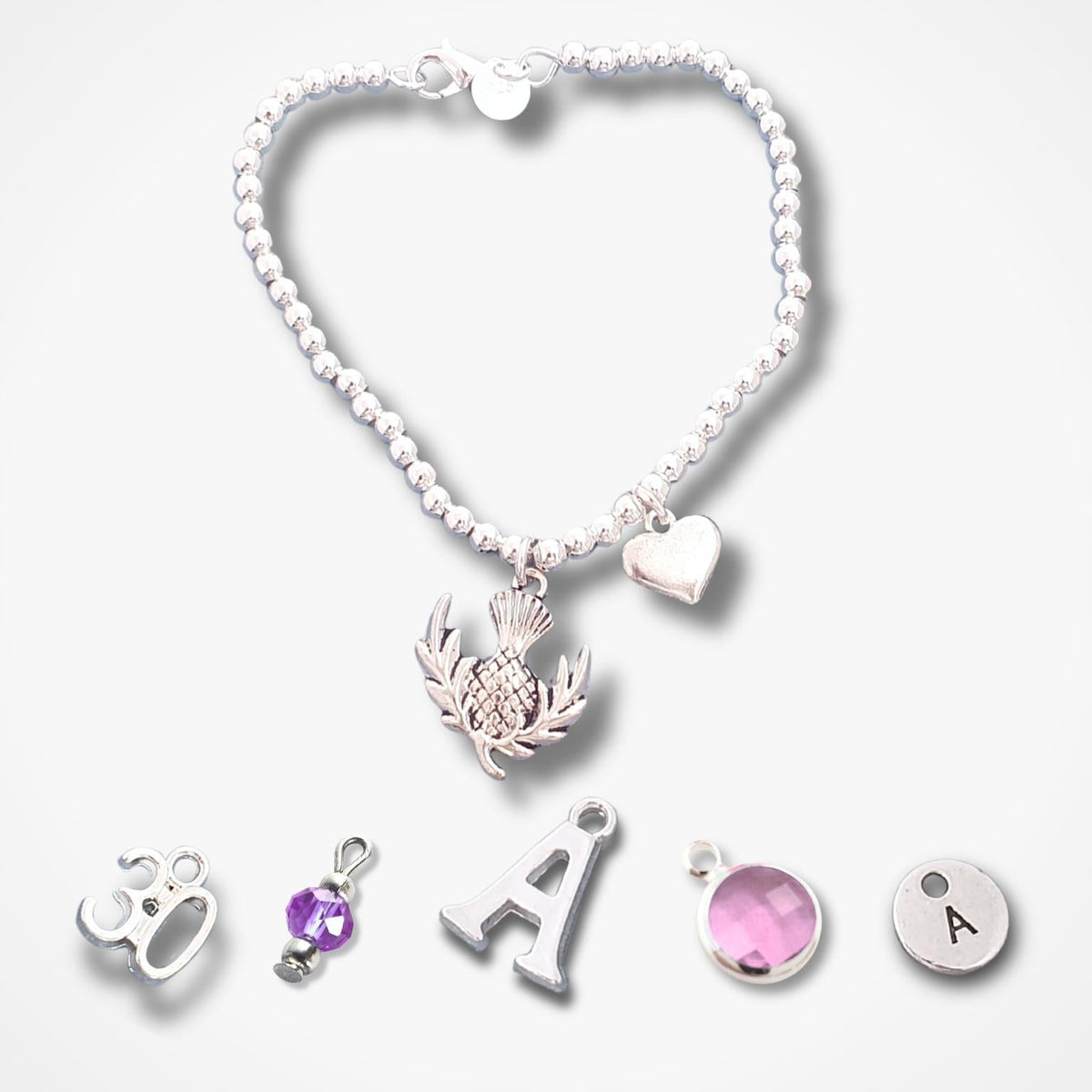 A silver beaded bracelet with a thistle pendant, a small heart charm, and five loose silver charms below it including a silver letter A, a round silver disc with the letter A, a pink gemstone charm, and a purple gemstone charm, all on a light grey background.