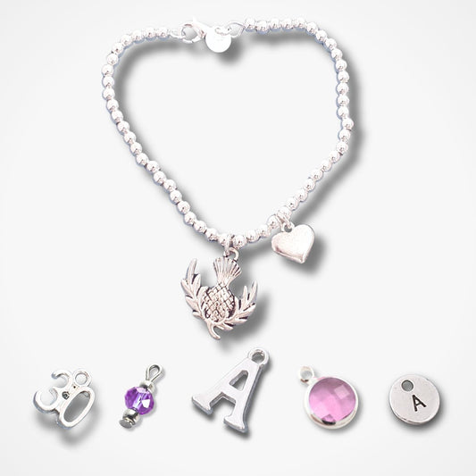 A silver beaded bracelet with a thistle pendant, a small heart charm, and five loose silver charms below it including a silver letter A, a round silver disc with the letter A, a pink gemstone charm, and a purple gemstone charm, all on a light grey background.
