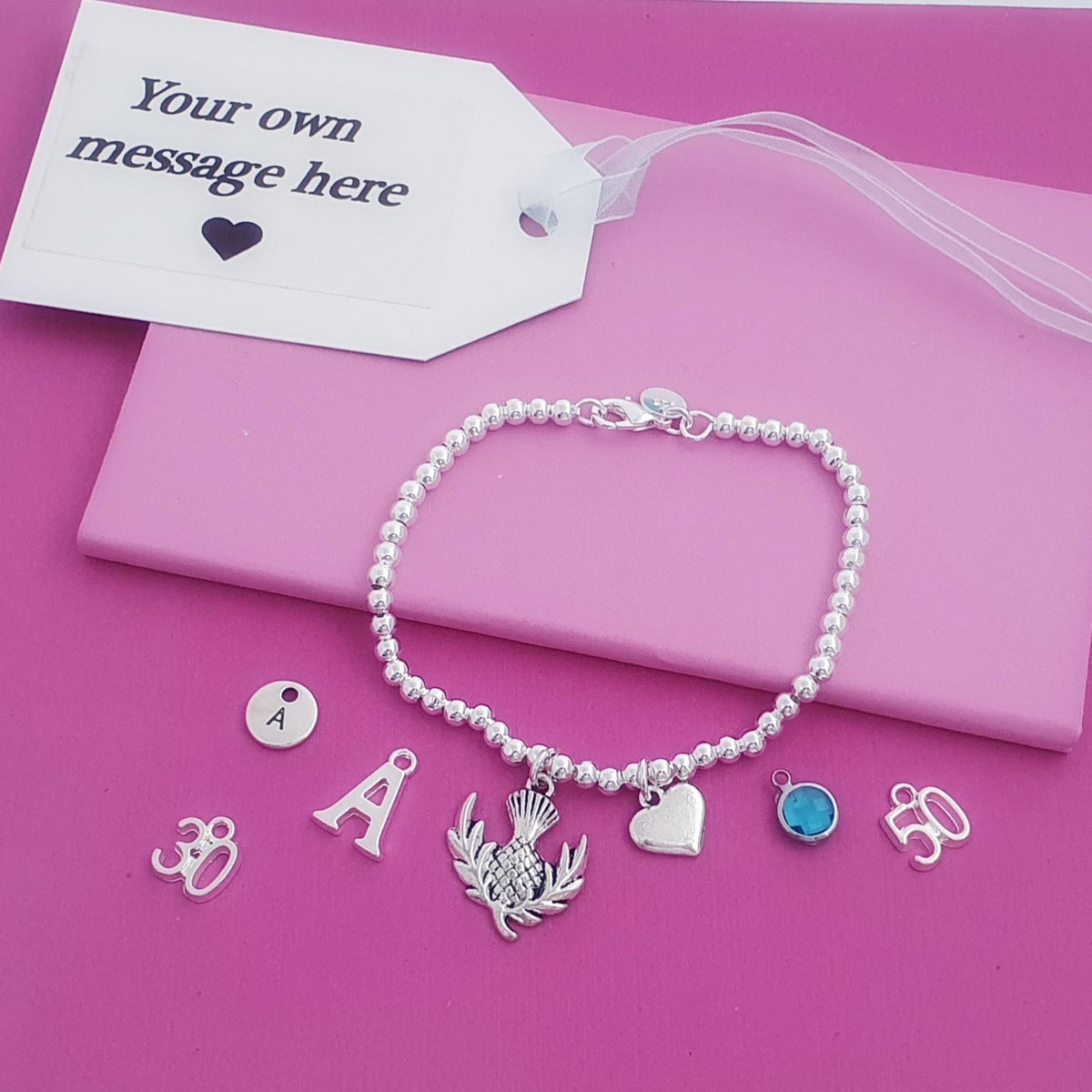 A silver beaded bracelet with multiple customizable charms, displayed on a pink gift box with a white message tag. The charms include a silver thistle (representing Scotland), a silver heart, a blue stone, and letters, set against a solid pink background.