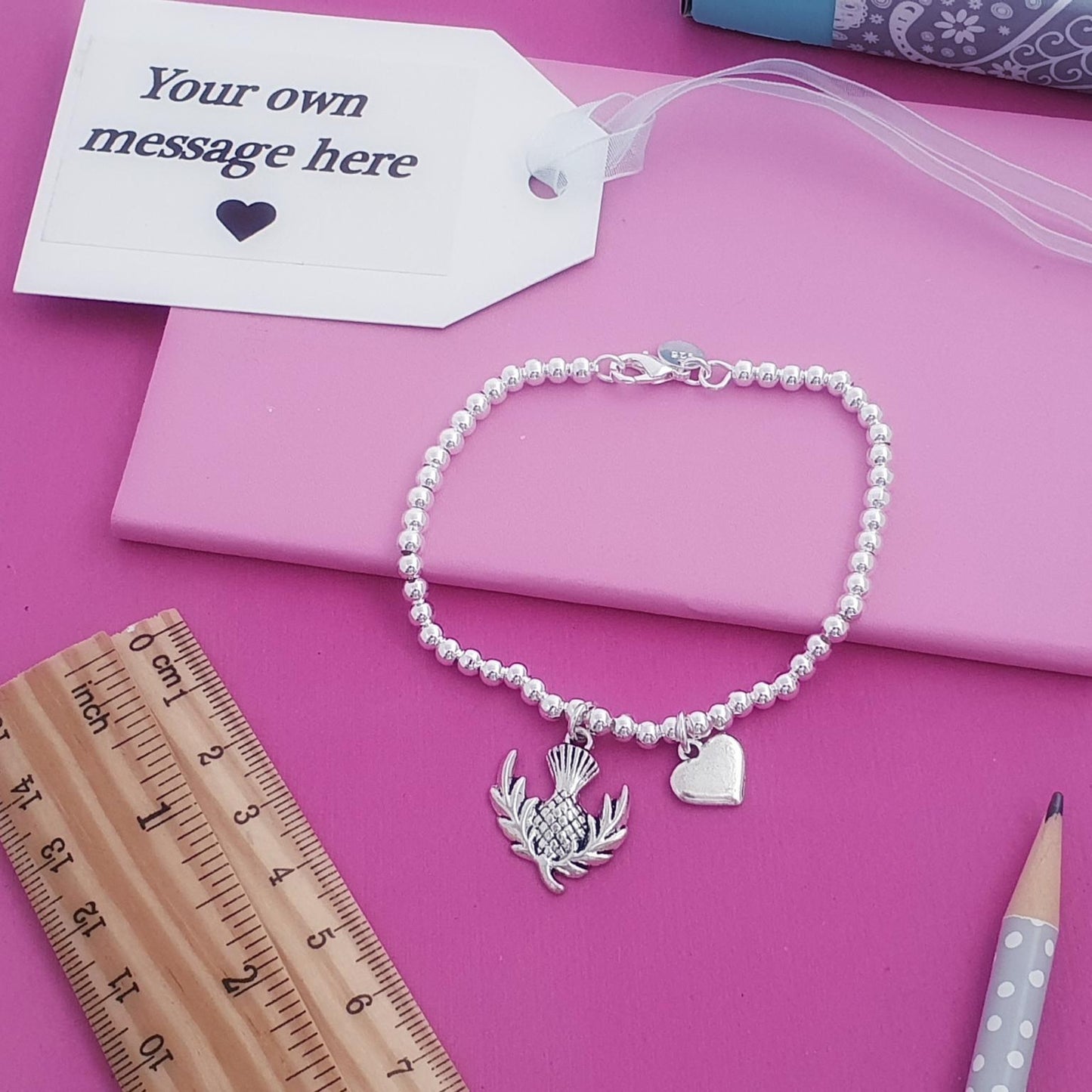 A silver beaded bracelet with a thistle and heart charm, displayed on a pink notebook with a white message tag, wooden ruler, and pencil on a pink background.