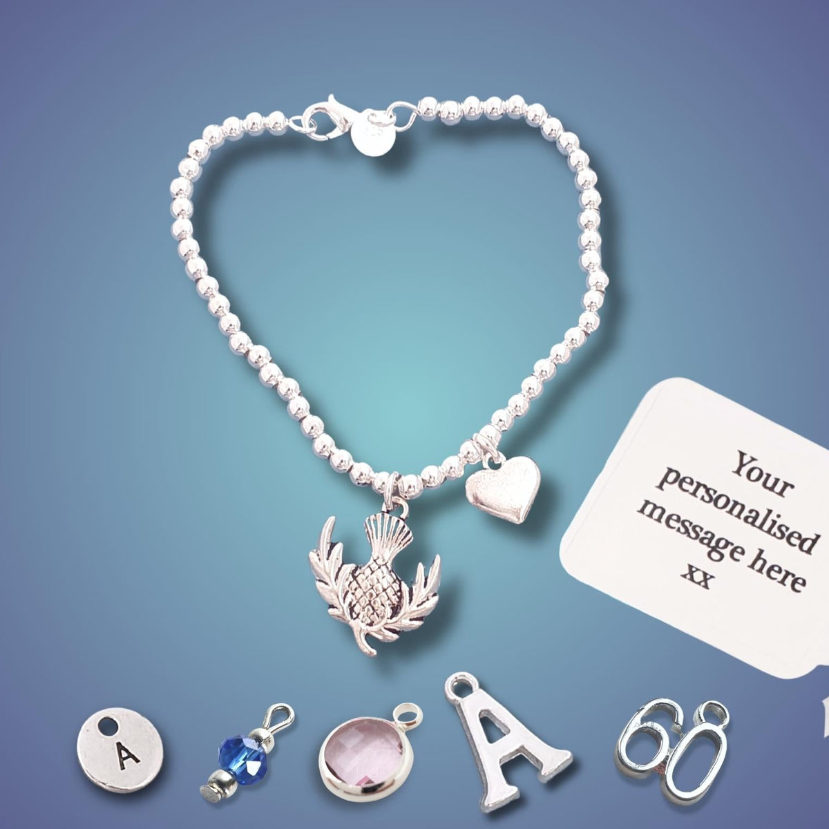 A silver bead bracelet with a silver thistle pendant and heart charm, displayed on a blue gradient background. The image includes small silver charms (letter A, blue bead, pink stone, letter A, number 66) and a white card that says 'Your personalised message here xx' with a hand holding the tail end of the card.