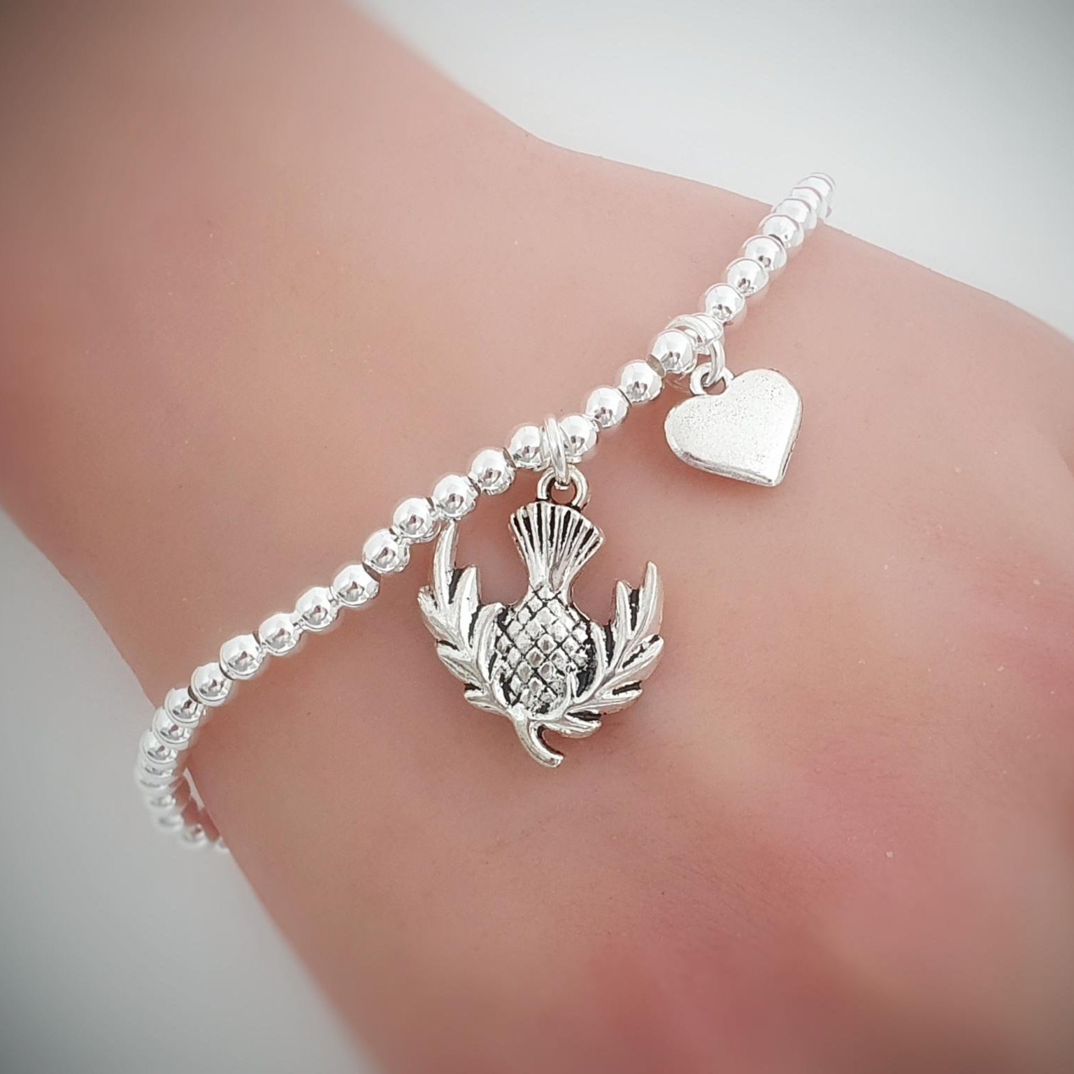 A close-up of a delicate silver beaded bracelet worn on a wrist. The bracelet features a silver thistle charm and a small silver heart charm, set against a soft, blurred skin background.