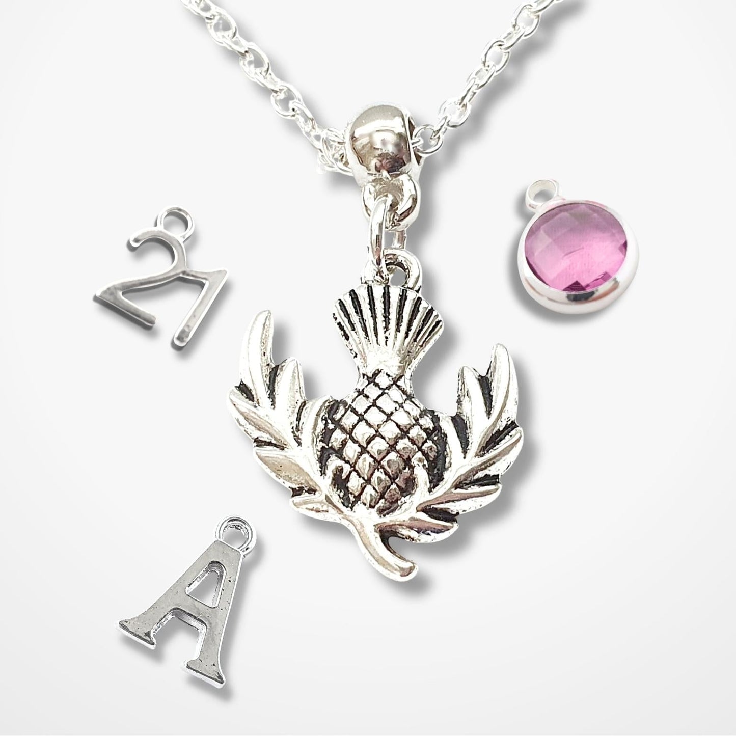 Silver necklace with thistle pendant, letter 'A' charm, and pink gemstone on a white background