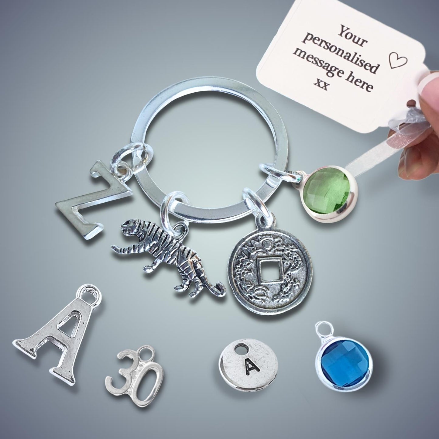 Silver keychain with tiger charm and coin charm and a hand holding a card for personalization on a gray background.