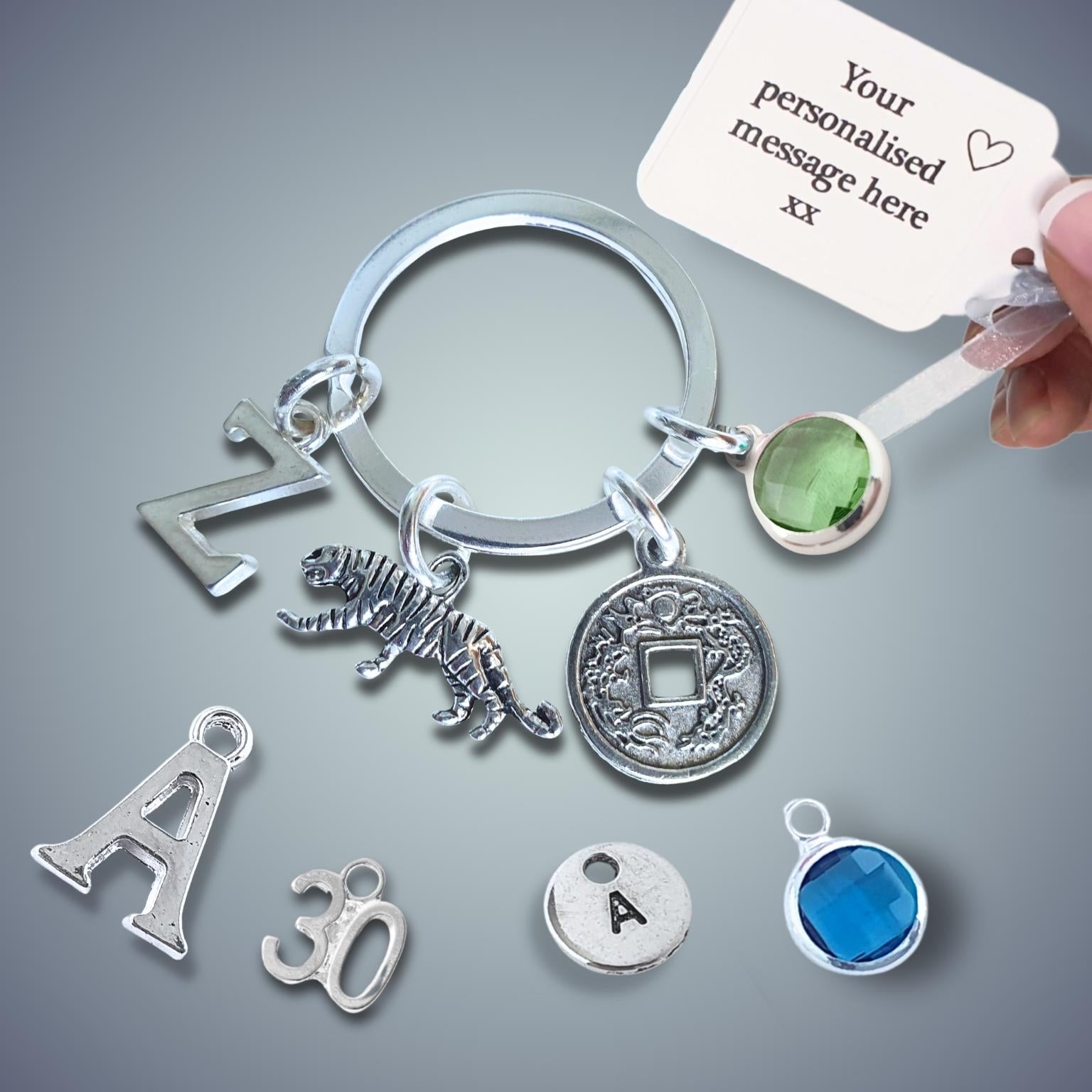 Silver keychain with tiger charm and coin charm and a hand holding a card for personalization on a gray background.