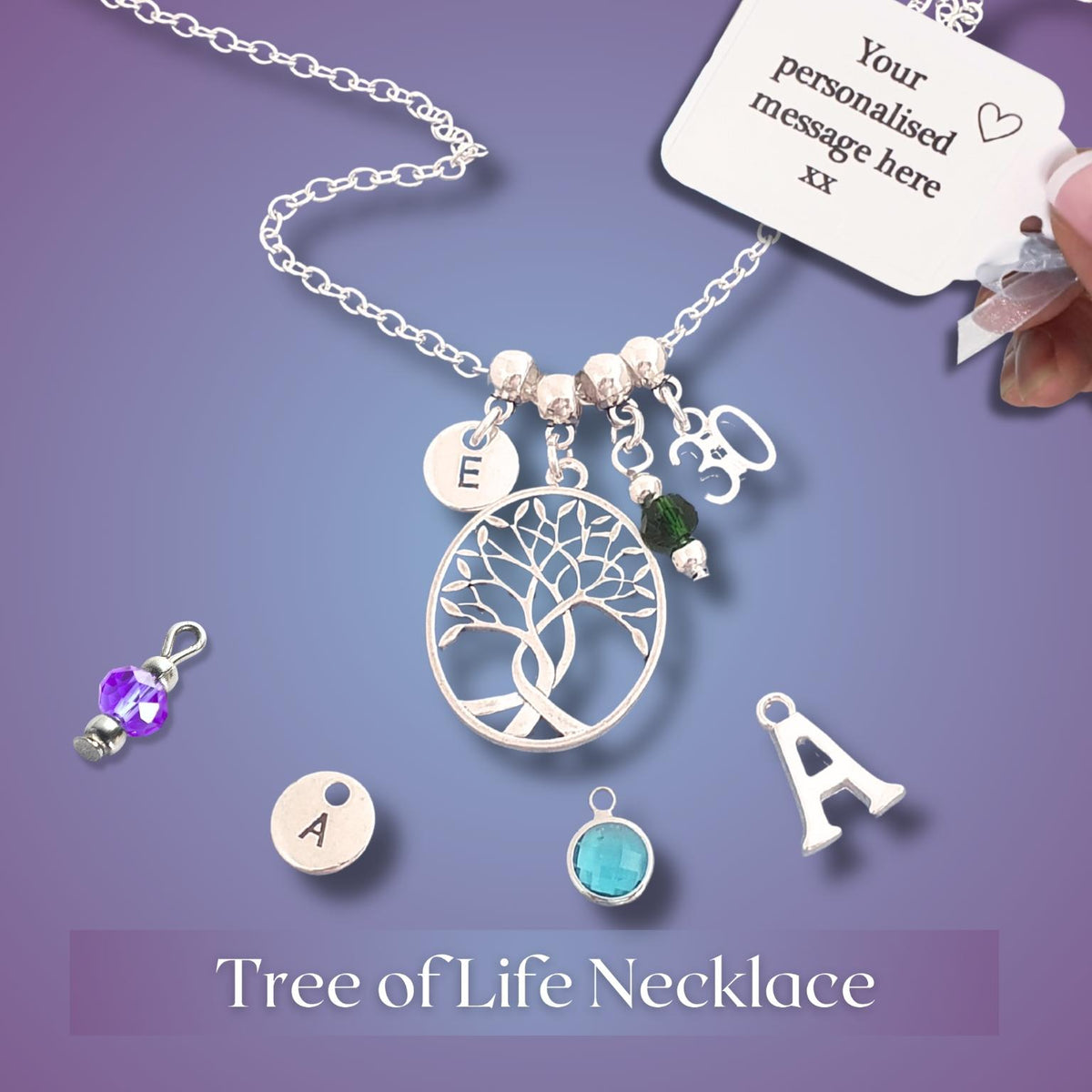 A silver tree of life charm necklace displayed on a purple gradient background, with a hand holding the attached white personalization card. The necklace features a circular tree of life pendant, a disc charm engraved with the letter 'E', a letter 'B' charm, a small green bead, and other small charms.