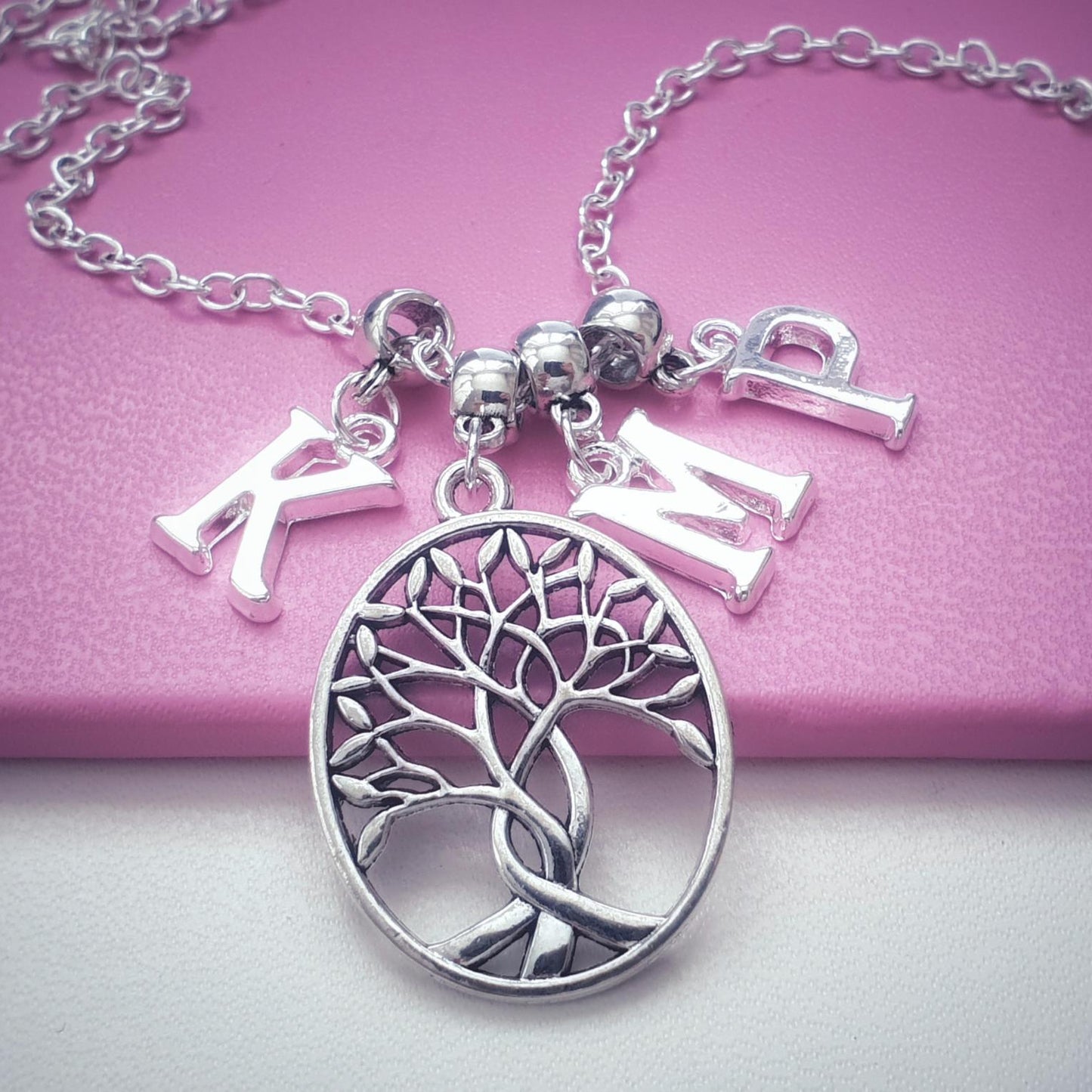 A silver necklace with a circular tree of life pendant and three dangling letter charms spelling 'KMP', displayed on a pink surface.