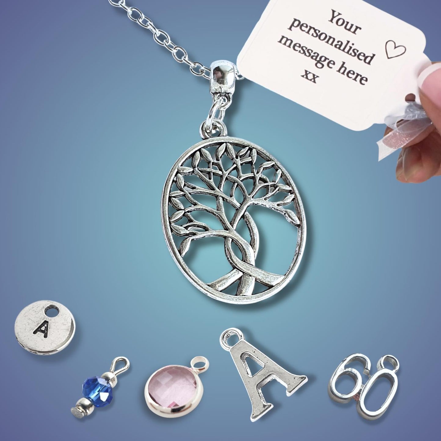 A silver tree of life pendant necklace on a chain, shown with a hand holding a customization tag. Several small silver charms and beads are arranged below on a blue gradient background.