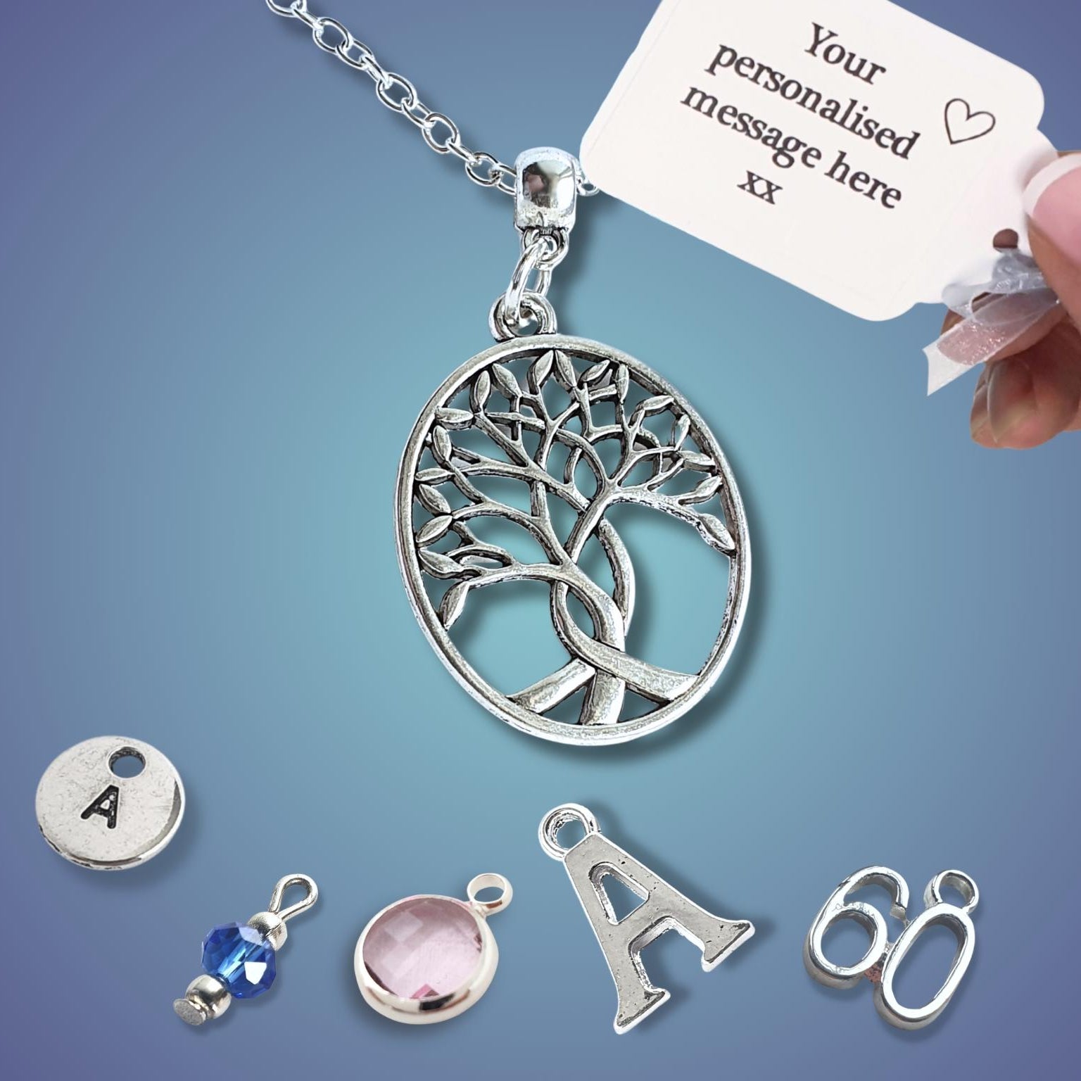 A silver tree of life pendant necklace on a chain, shown with a hand holding a customization tag. Several small silver charms and beads are arranged below on a blue gradient background.