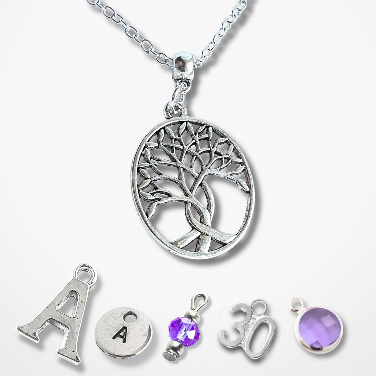 A silver Tree of Life pendant necklace displayed with five matching customizable charm options below it on a light gray background. The main pendant features a detailed tree design inside a circle, while the options below include a letter 'A', a letter 'A' with a gem, a purple gem charm, a '30' charm, and a purple gem bezel.