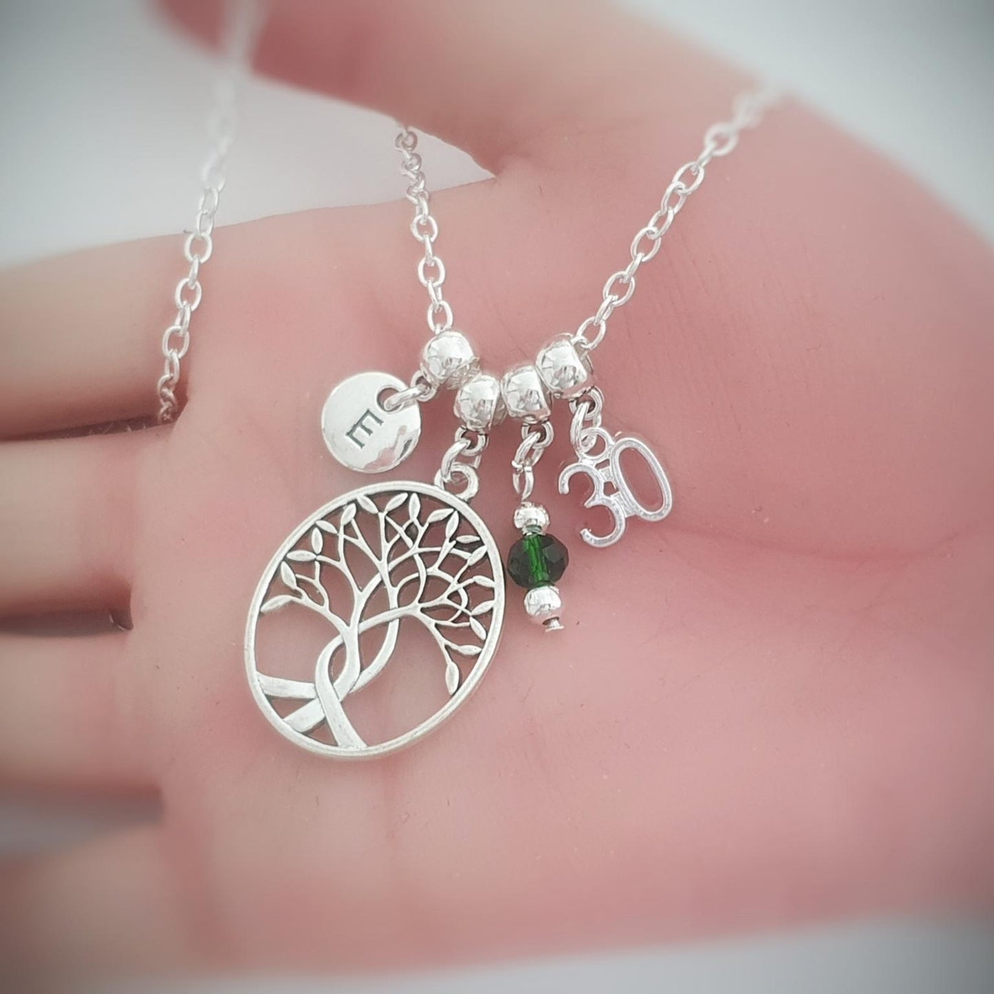 A silver tree of life pendant necklace with a tree design, a small Om symbol charm, a green heart-shaped bead, and a round engraved disc charm, displayed on a person's hand.