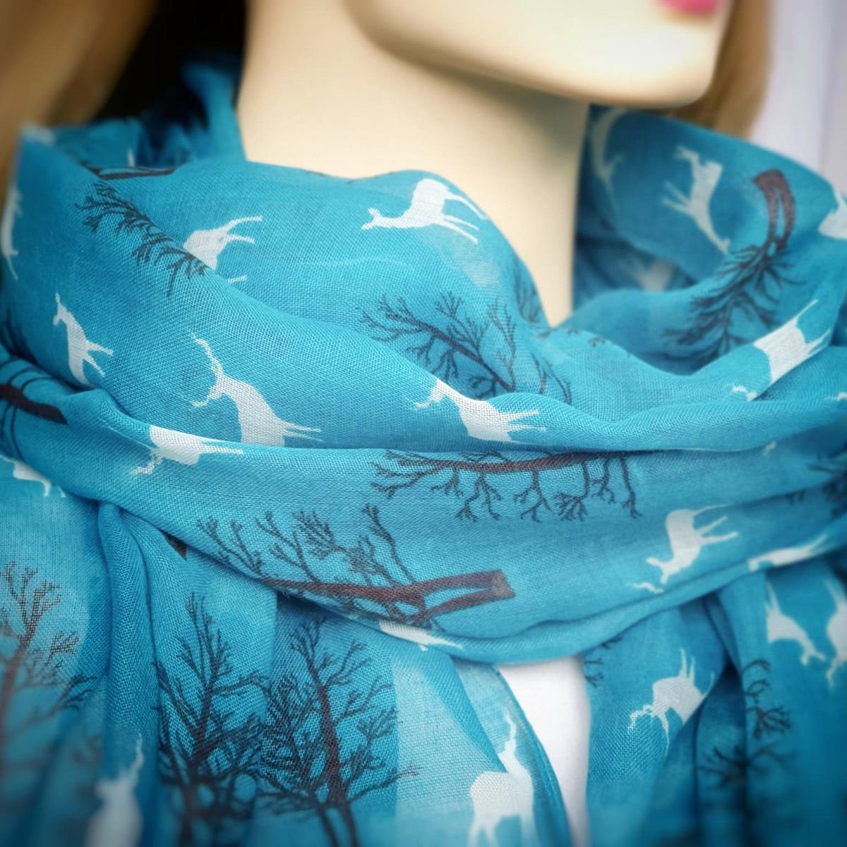 A close-up of a blue scarf with a pattern of white deer and dark grey tree branches, draped on a mannequin.