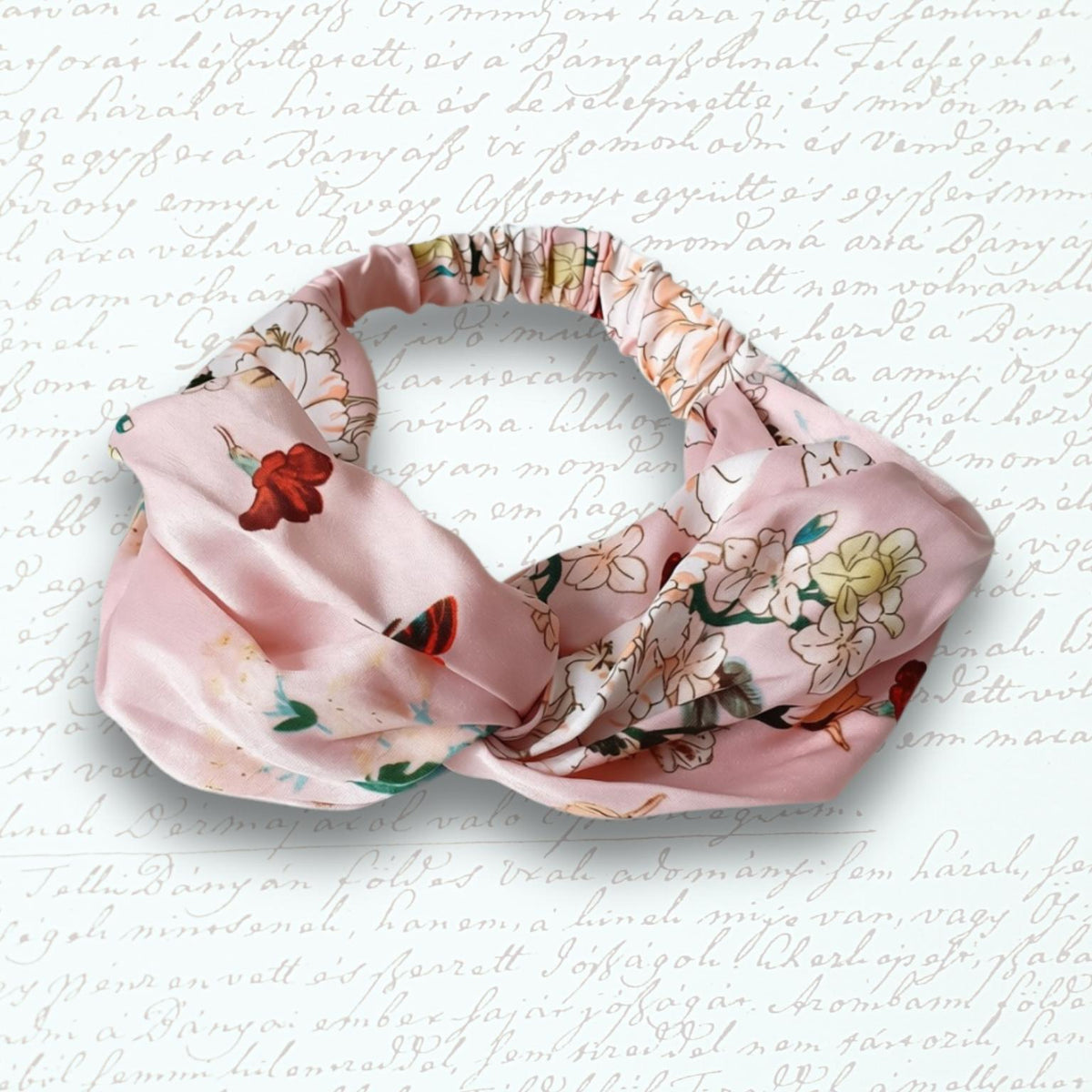 A pink satin headband with a knotted twist and a floral print, displayed on a textured white background with faint cursive writing.