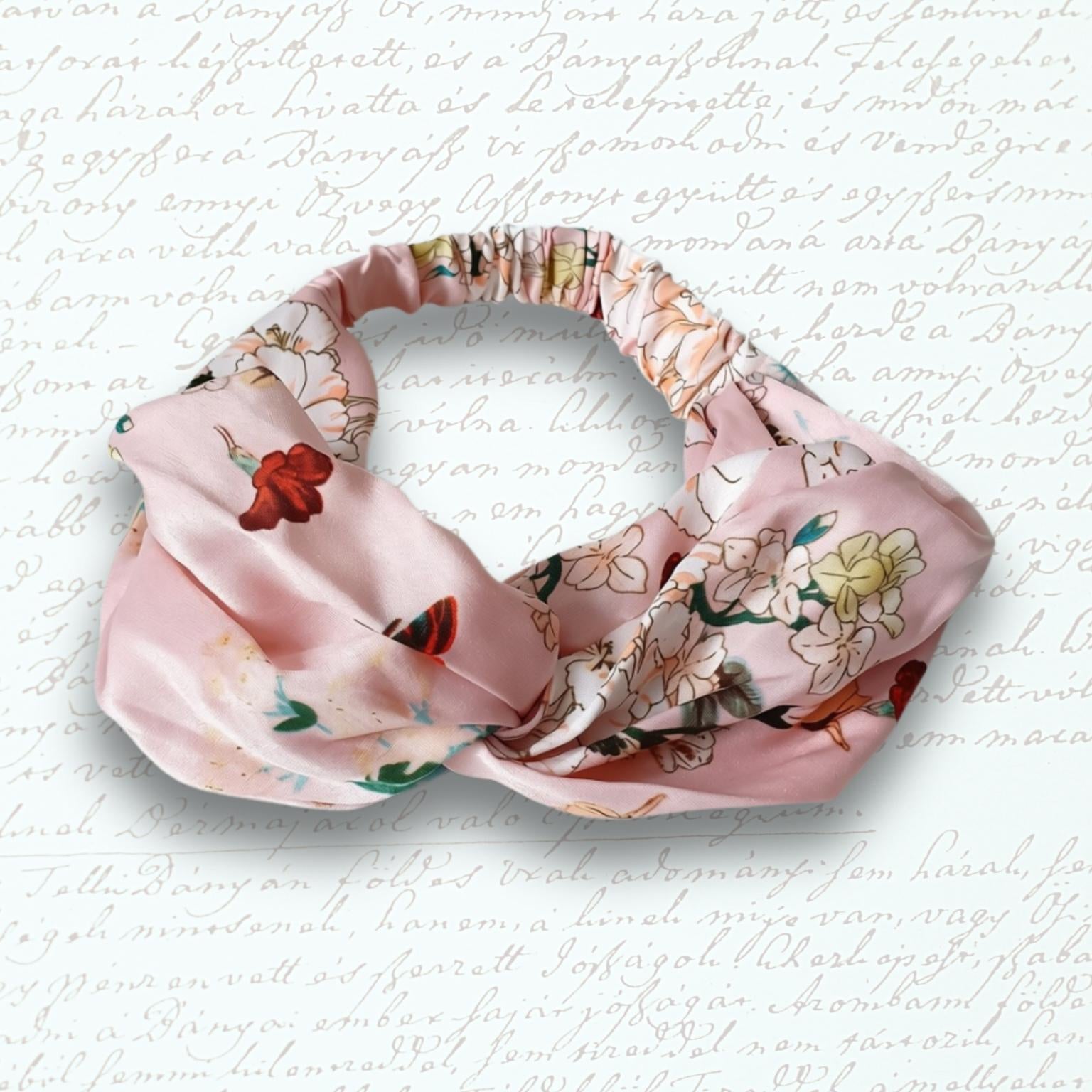 A pink satin headband with a knotted twist and a floral print, displayed on a textured white background with faint cursive writing.