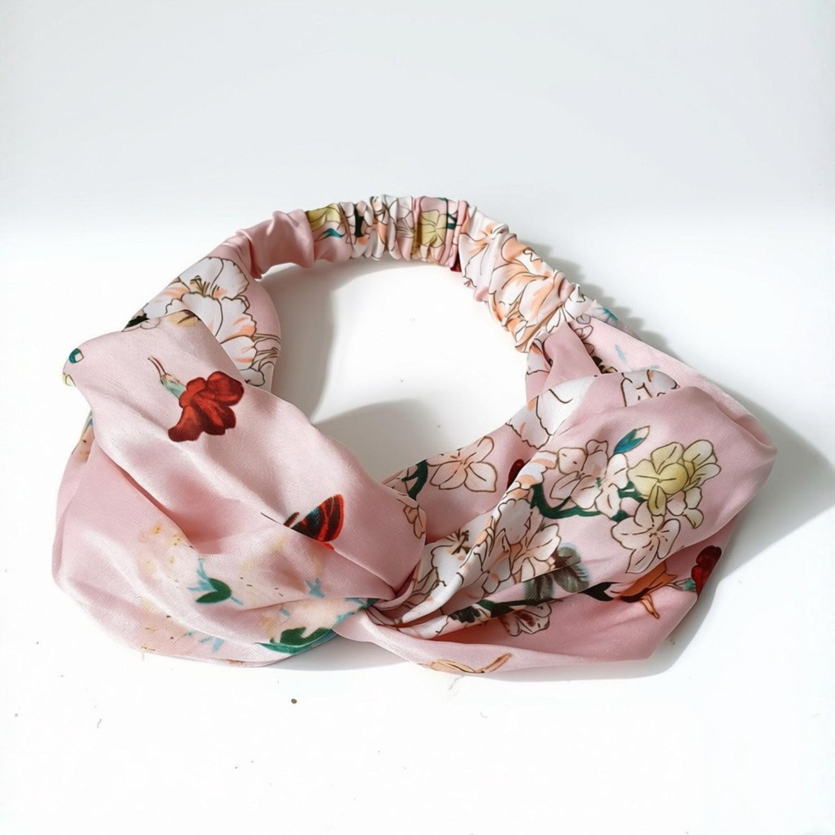A pink satin-like headband with a gathered elastic top and a floral print featuring white, red, and green flowers, displayed on a white background.