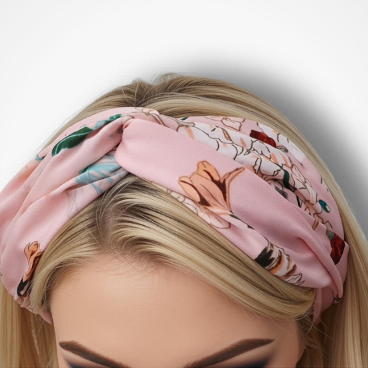 A close-up of a woman wearing a light pink silk headband with a botanical and floral print, styled in a knot on her head against a plain white background.