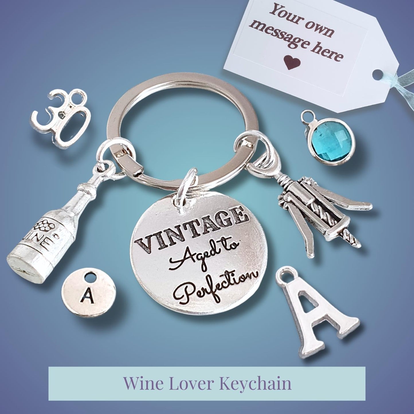 A silver-tone wine-themed keychain with charms including a bottle, corkscrew, wine glass, letter 'A', and a round engraved disc reading 'VINTAGE Aged to Perfection'. A small blue gem charm and a white customizable tag are shown on a blue gradient background.
