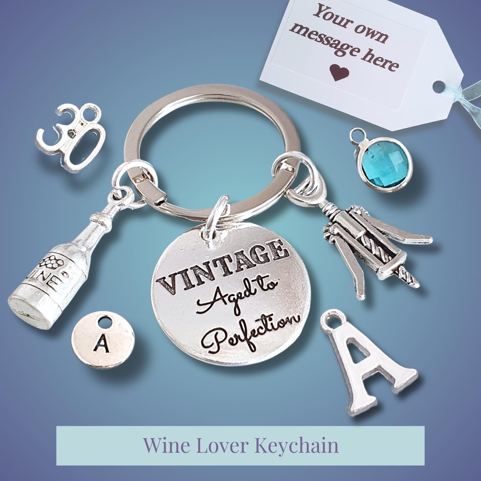 A silver-tone wine-themed keychain with charms including a bottle, corkscrew, wine glass, letter 'A', and a round engraved disc reading 'VINTAGE Aged to Perfection'. A small blue gem charm and a white customizable tag are shown on a blue gradient background.