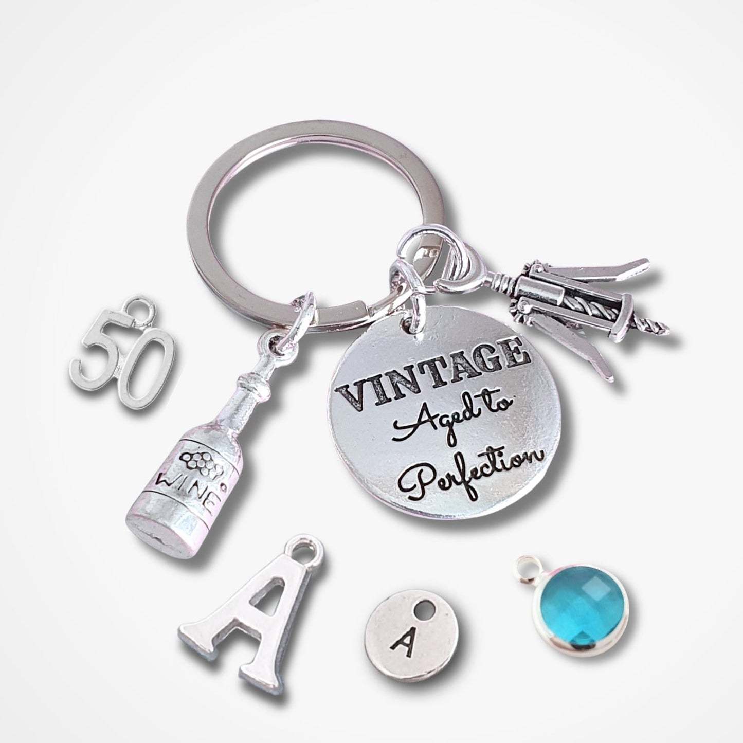 A silver-tone keychain with multiple charms, including a round disc engraved 'VINTAGE Aged To Perfection,' a bottle charm, a '50' charm, an 'A' letter charm, a small round 'A' charm, and a faceted blue gemstone charm, shown on a light gray background.
